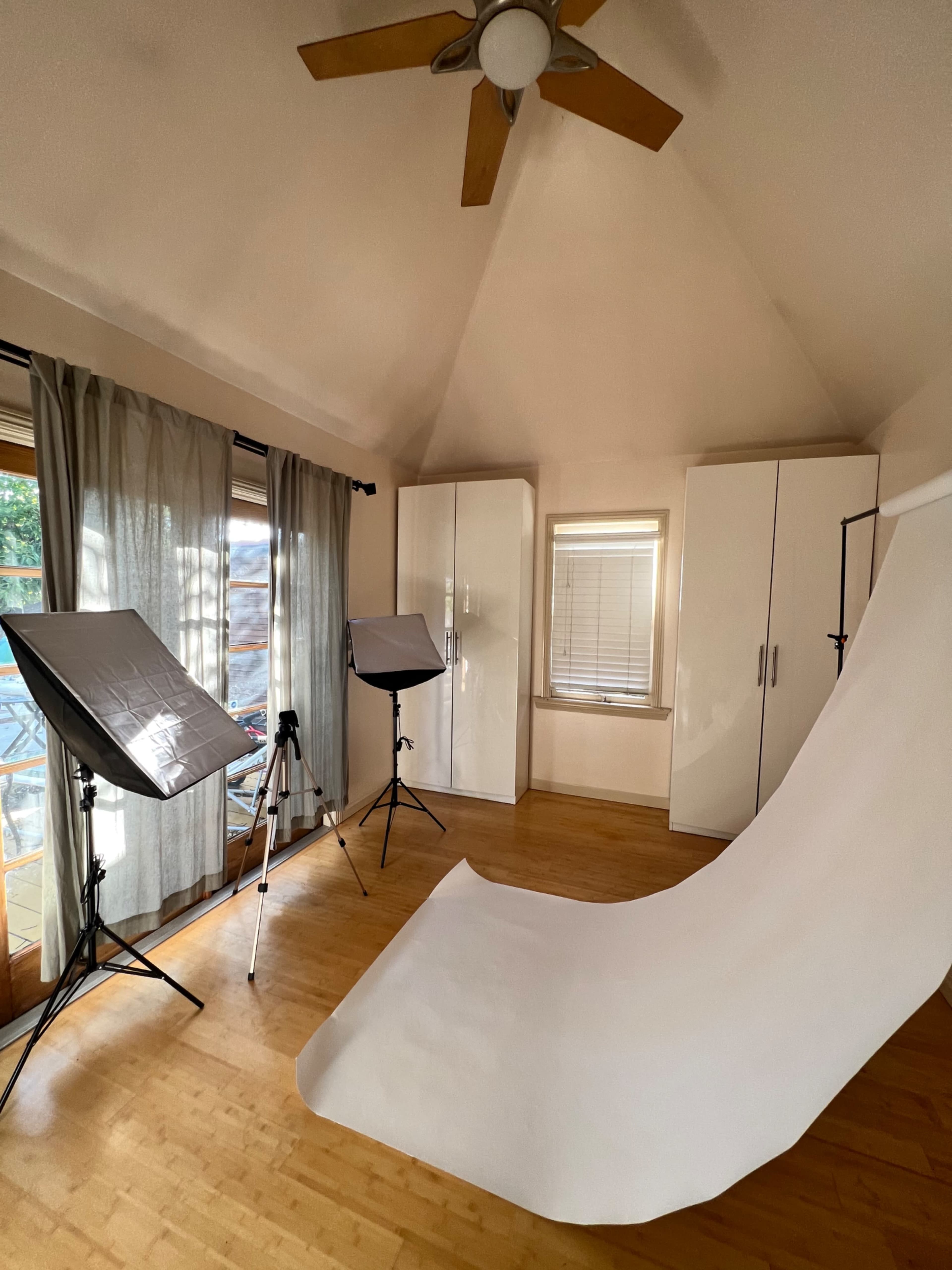 The room features a smooth, white backdrop on the floor, two softbox lights positioned on either side, and a tripod, with large windows providing natural light.