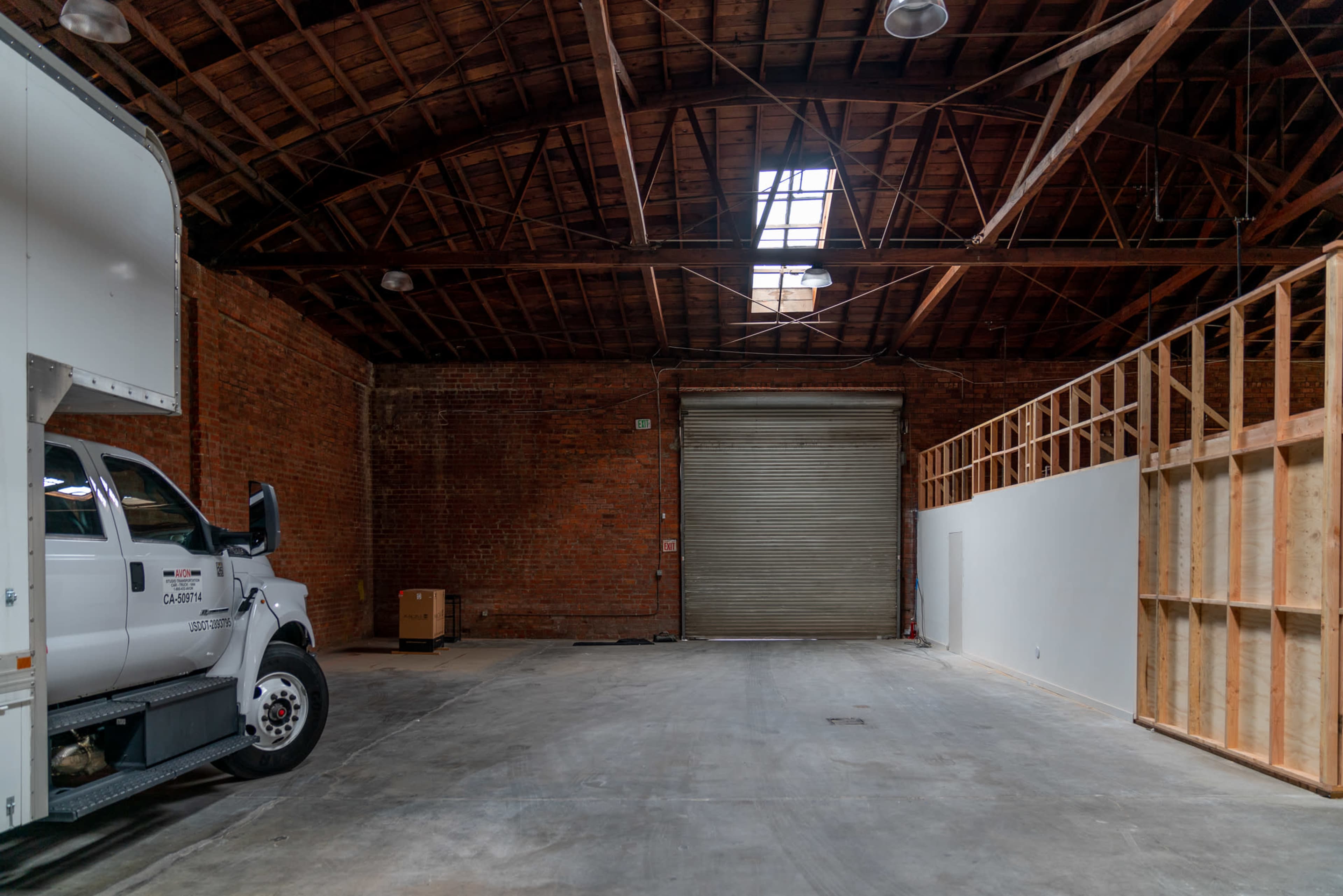 A spacious industrial warehouse features a truck parked beside a large roll-up door and exposed wooden beams overhead.
