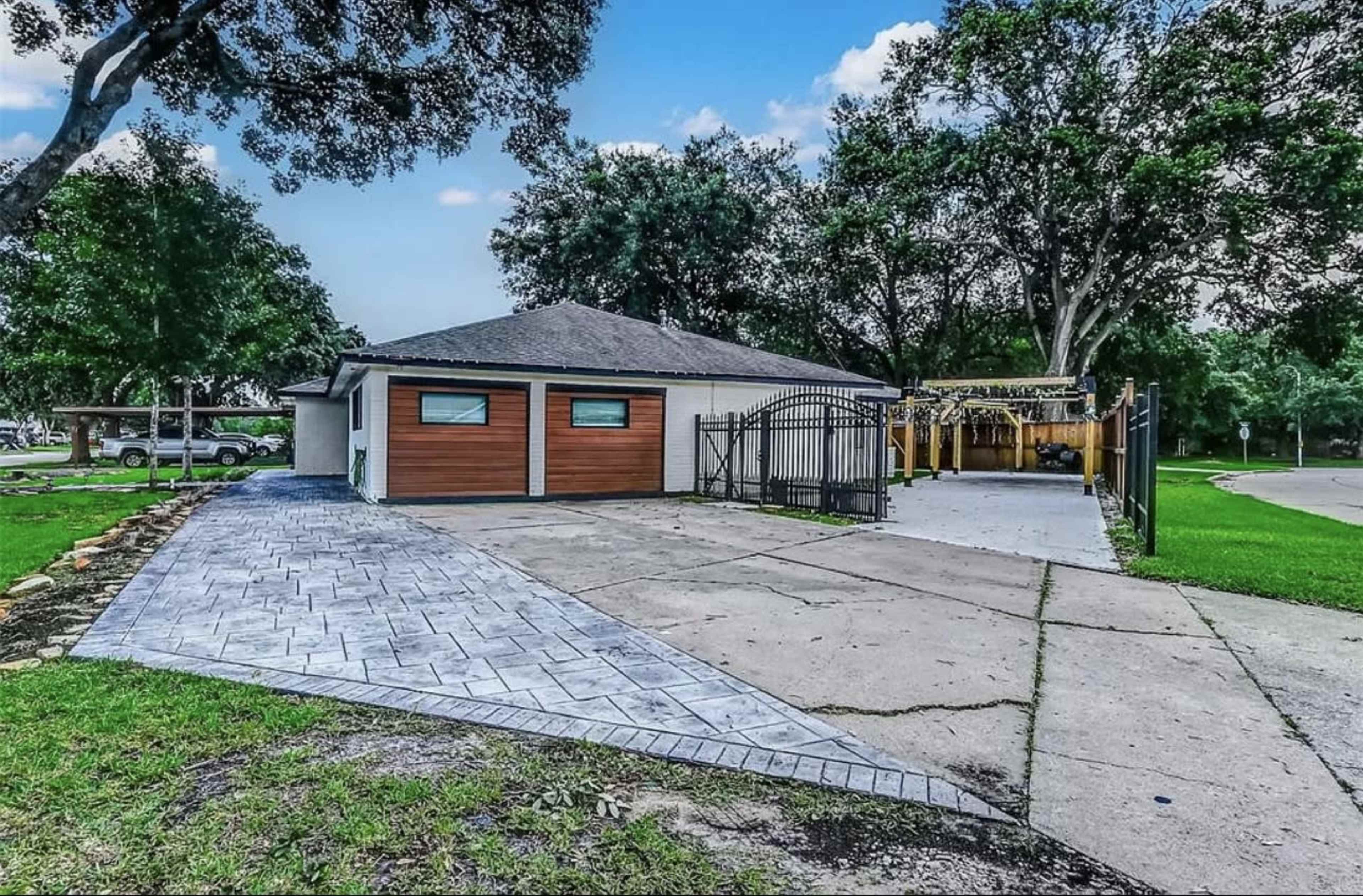The image shows a modern house with a driveway leading to a gated patio area, surrounded by greenery and trees.
