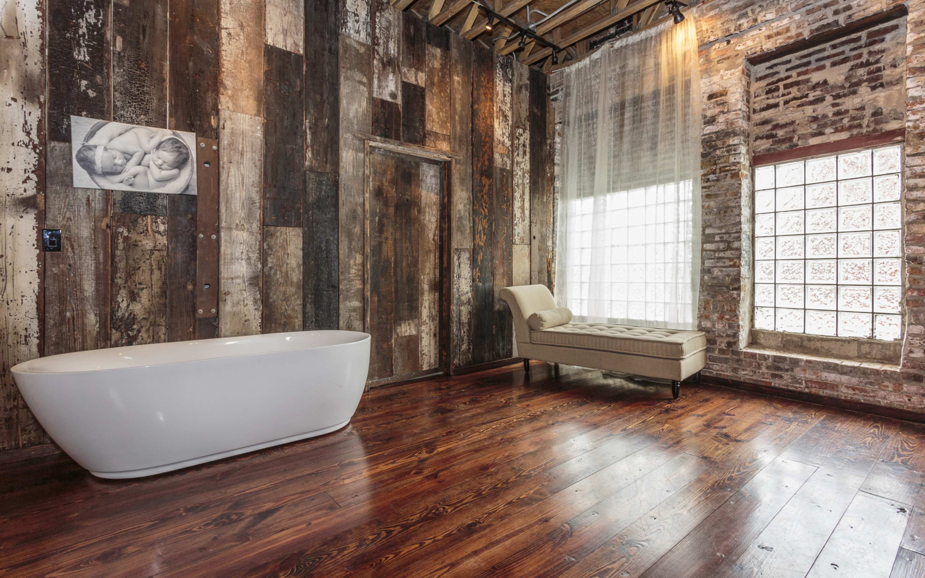 A modern bathroom with a freestanding white bathtub, a chaise lounge, exposed brick and wooden walls, and large windows allowing natural light to enter.