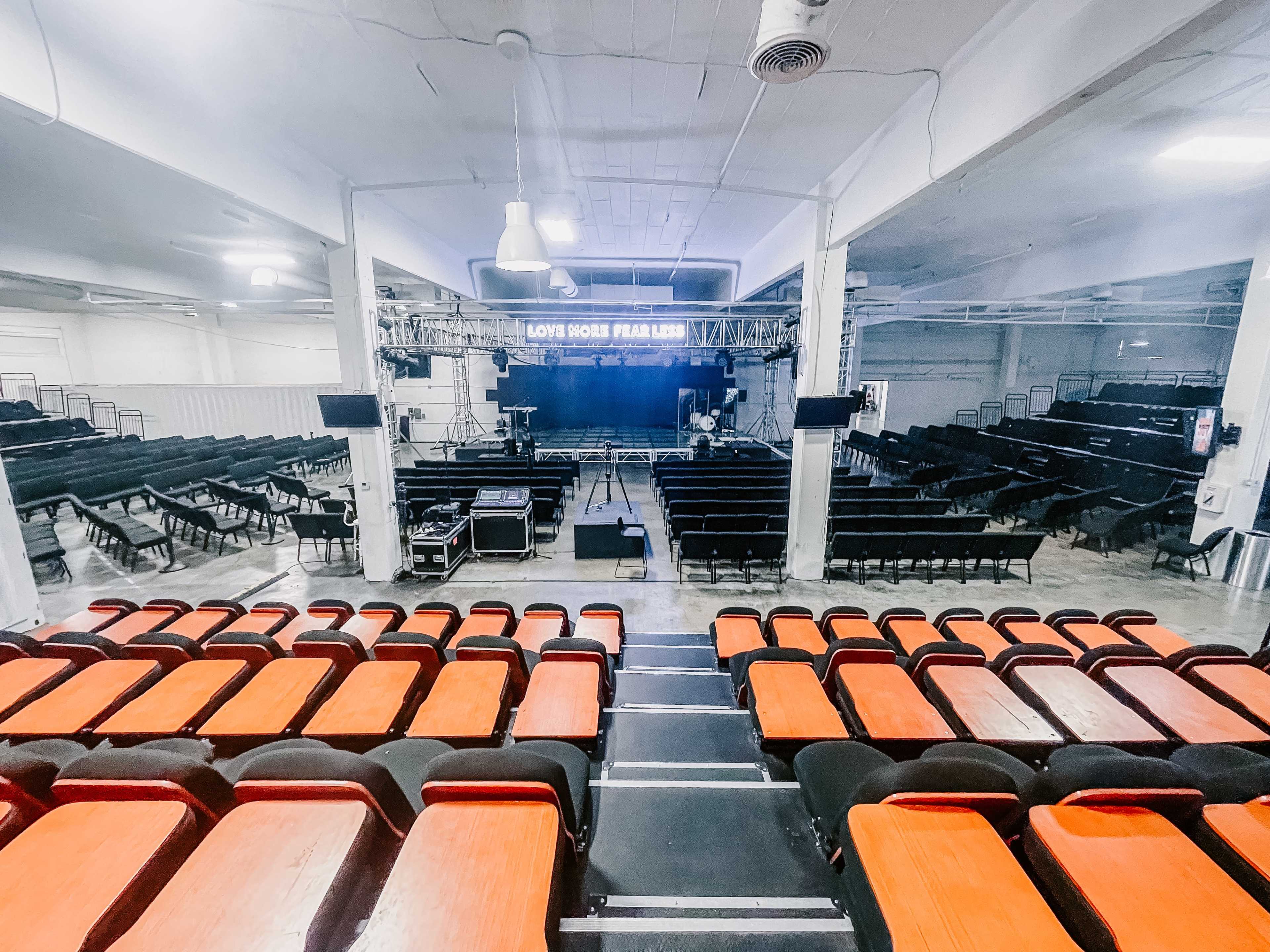 An empty auditorium with rows of red and black seats facing a stage equipped with lighting and sound equipment.