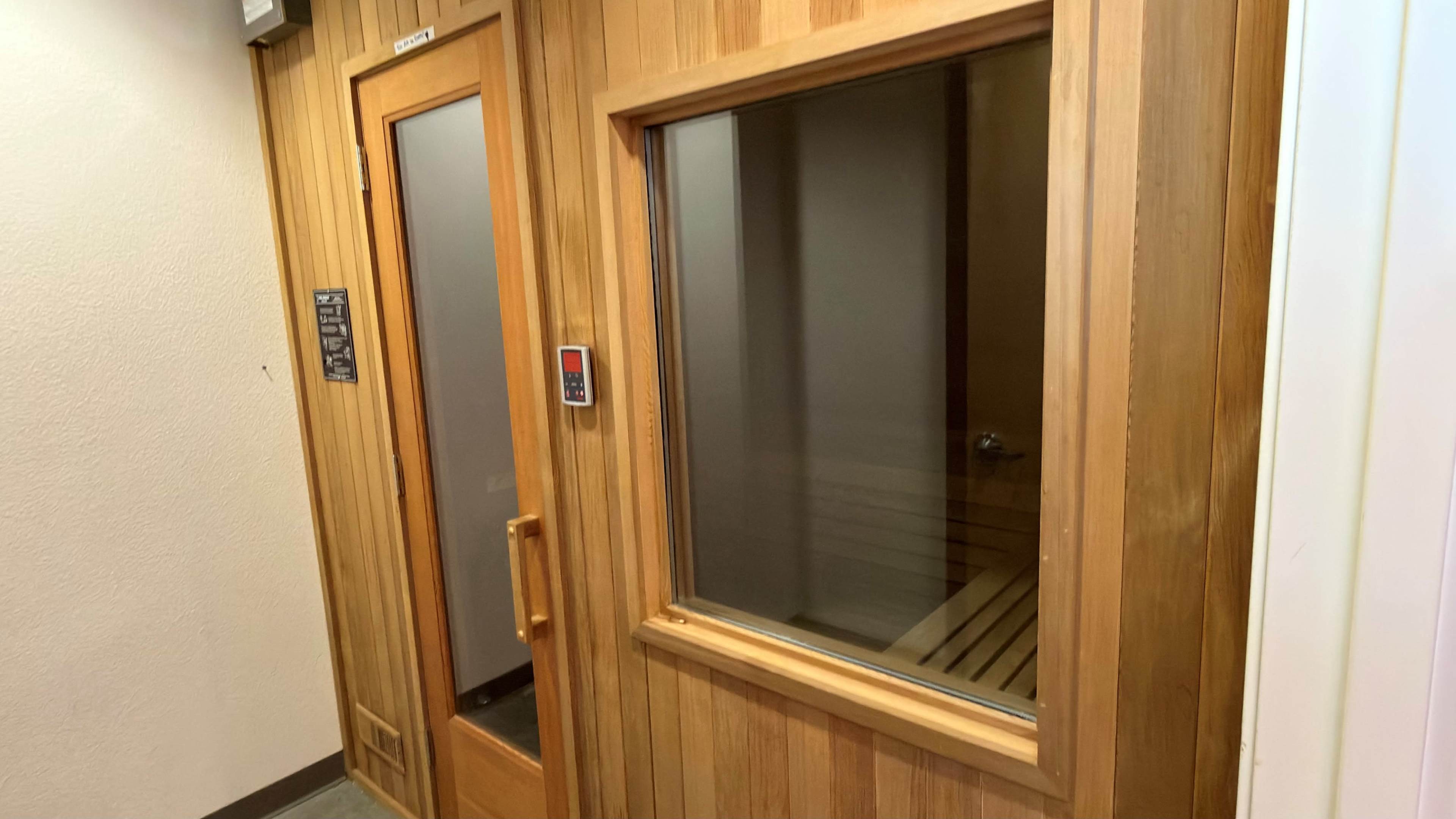 The image features a wooden sauna room entrance with large glass panels and a digital control panel mounted on the wall.