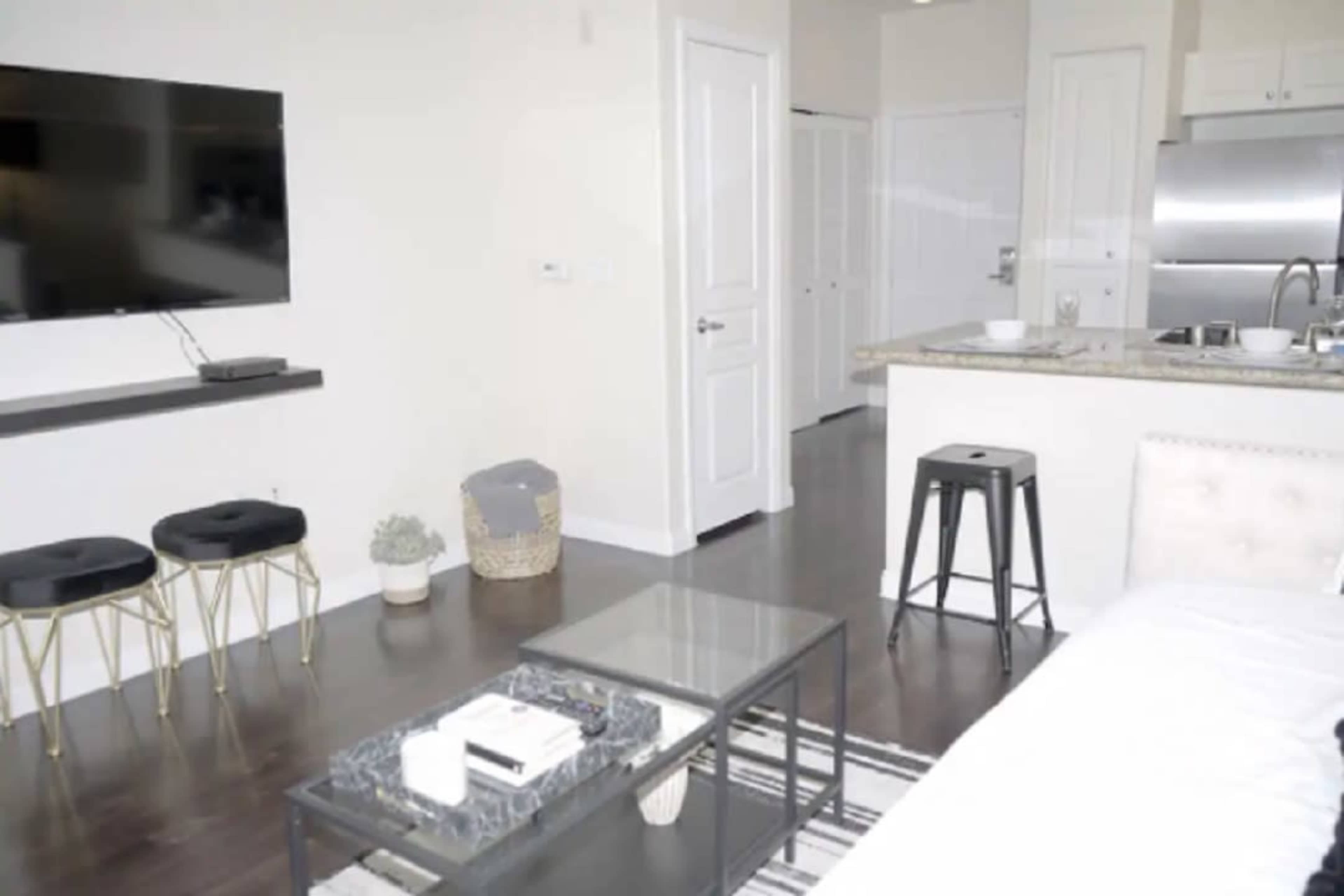 The image shows a modern living space with a television mounted on the wall, a small kitchen area with a bar counter, and minimalist furniture including two stools and a coffee table.