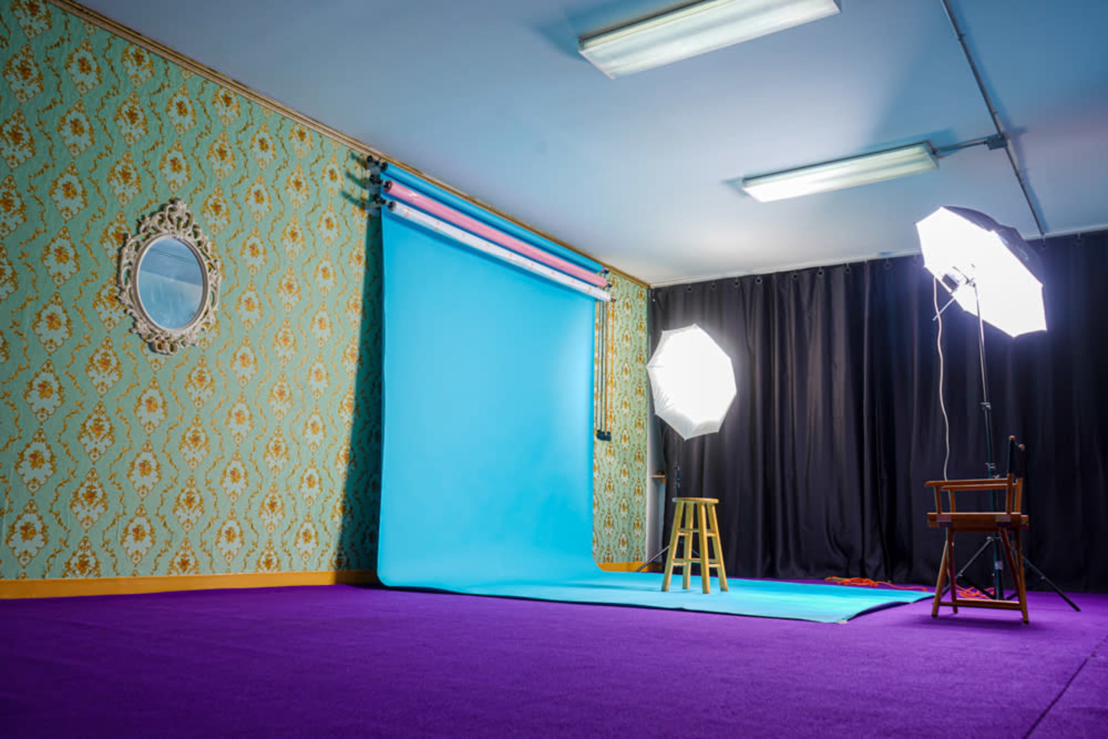 A photo studio is set up with a blue backdrop, two softbox lights, a stool, and a chair against patterned wallpaper and a purple carpet.
