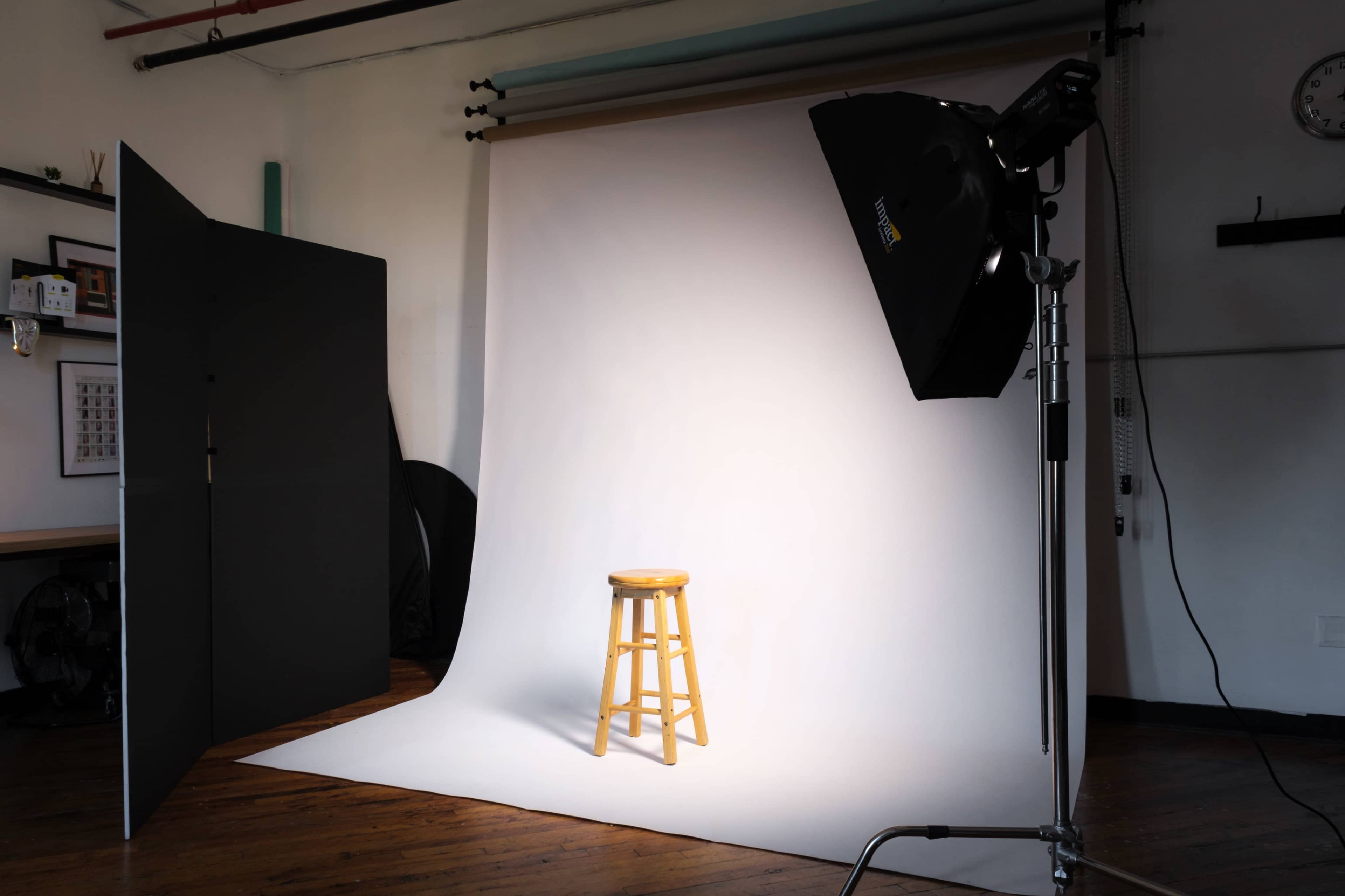 A wooden stool is positioned under a spotlight in a photography studio with a plain backdrop.