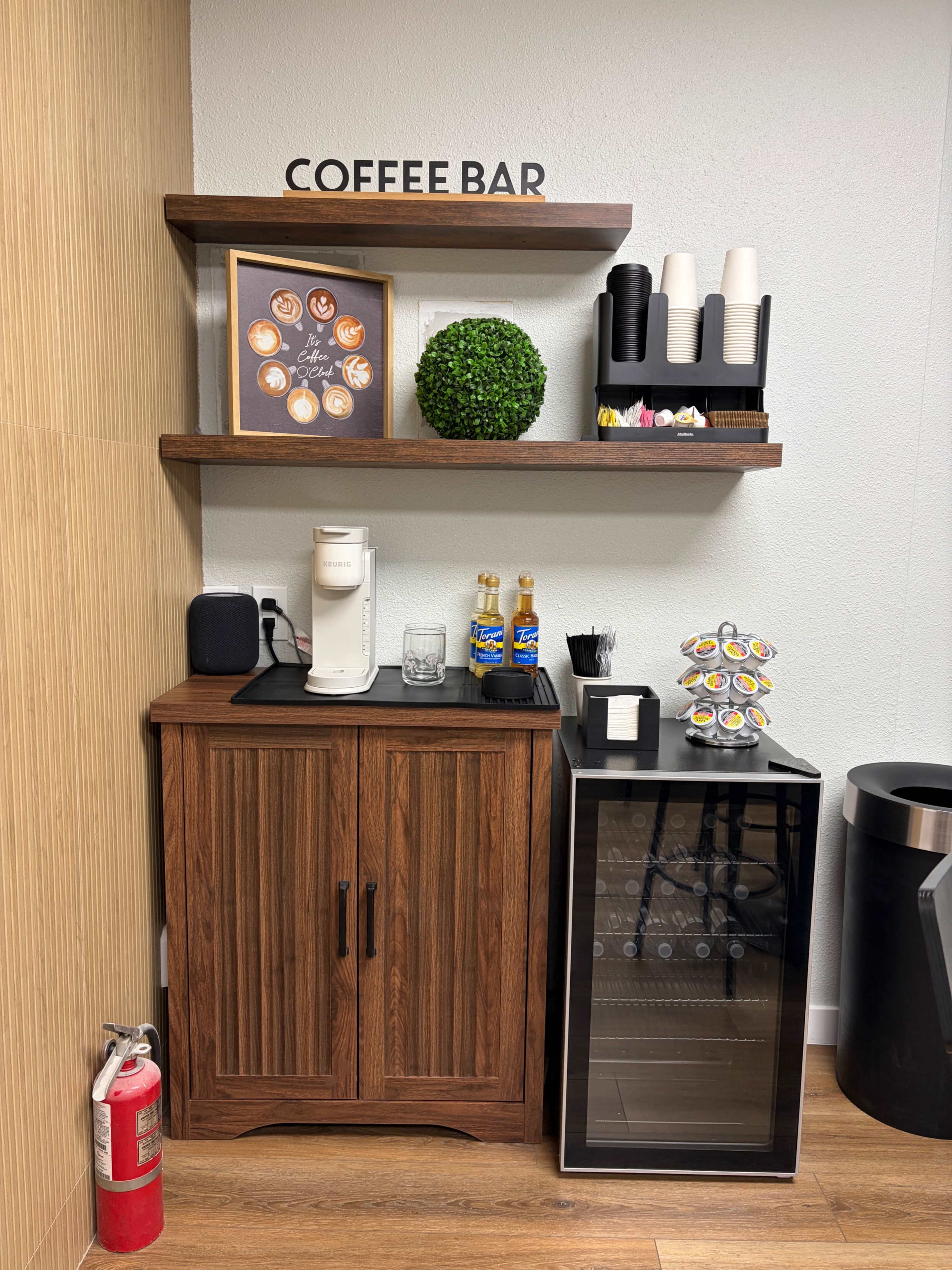 The image shows a coffee bar featuring a wooden cabinet with a countertop, a coffee machine, and a small fridge, accompanied by shelves above displaying decorative items and coffee supplies.