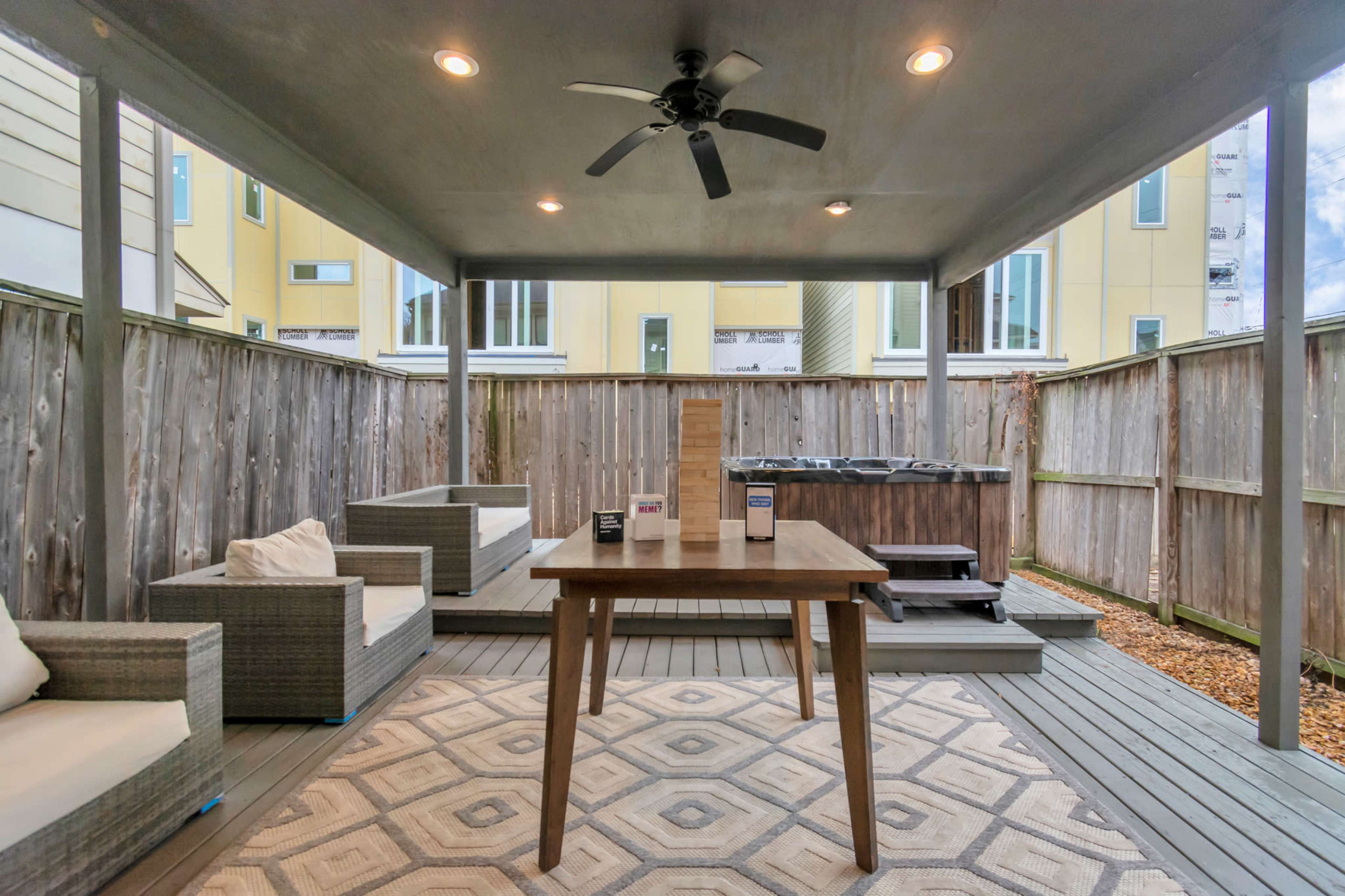 The image shows a covered outdoor patio with a wooden table, two seating areas, and a hot tub against a wooden fence.