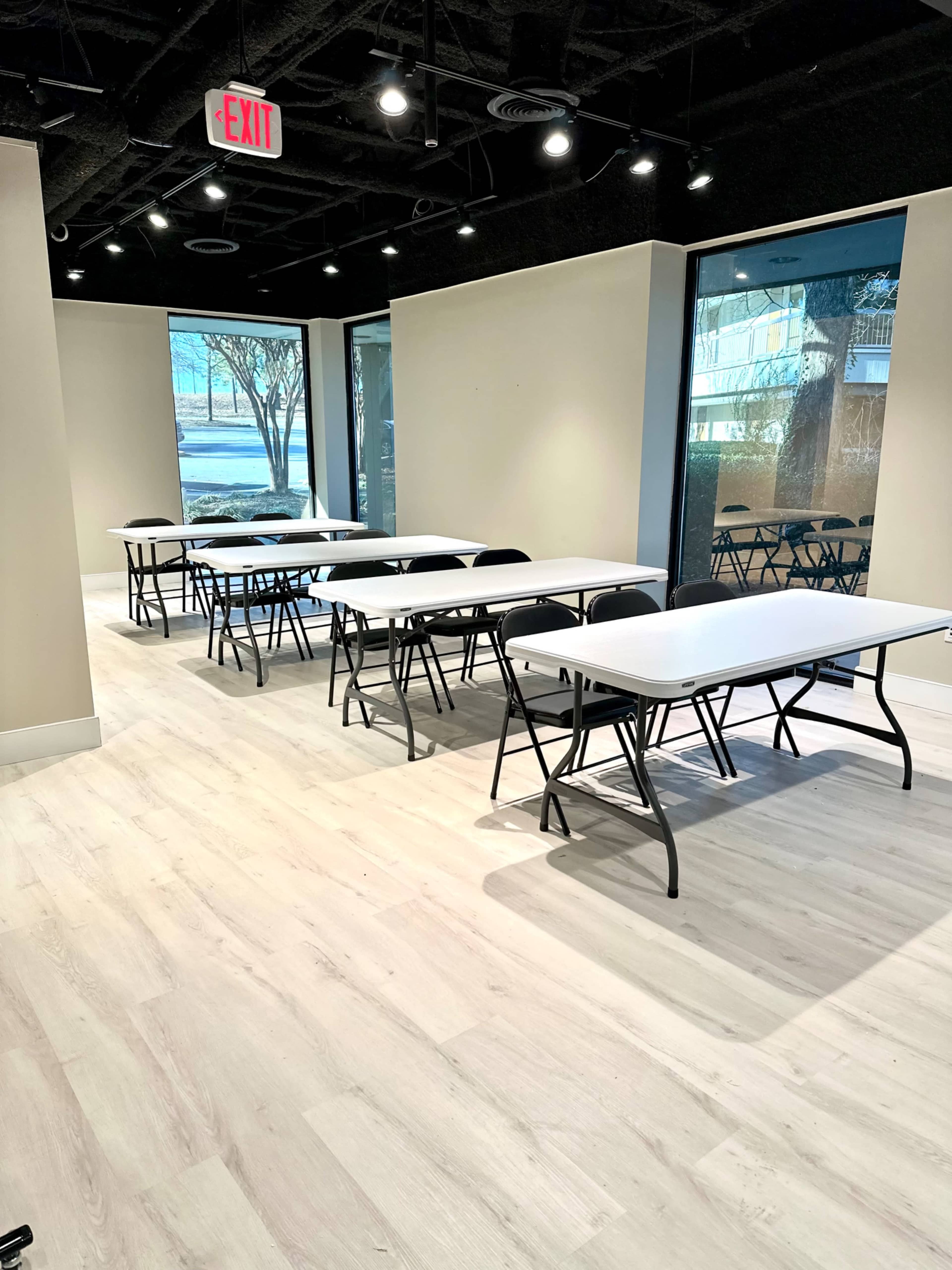 A spacious room with several rows of folding tables and chairs arranged neatly, illuminated by natural light coming through large windows.