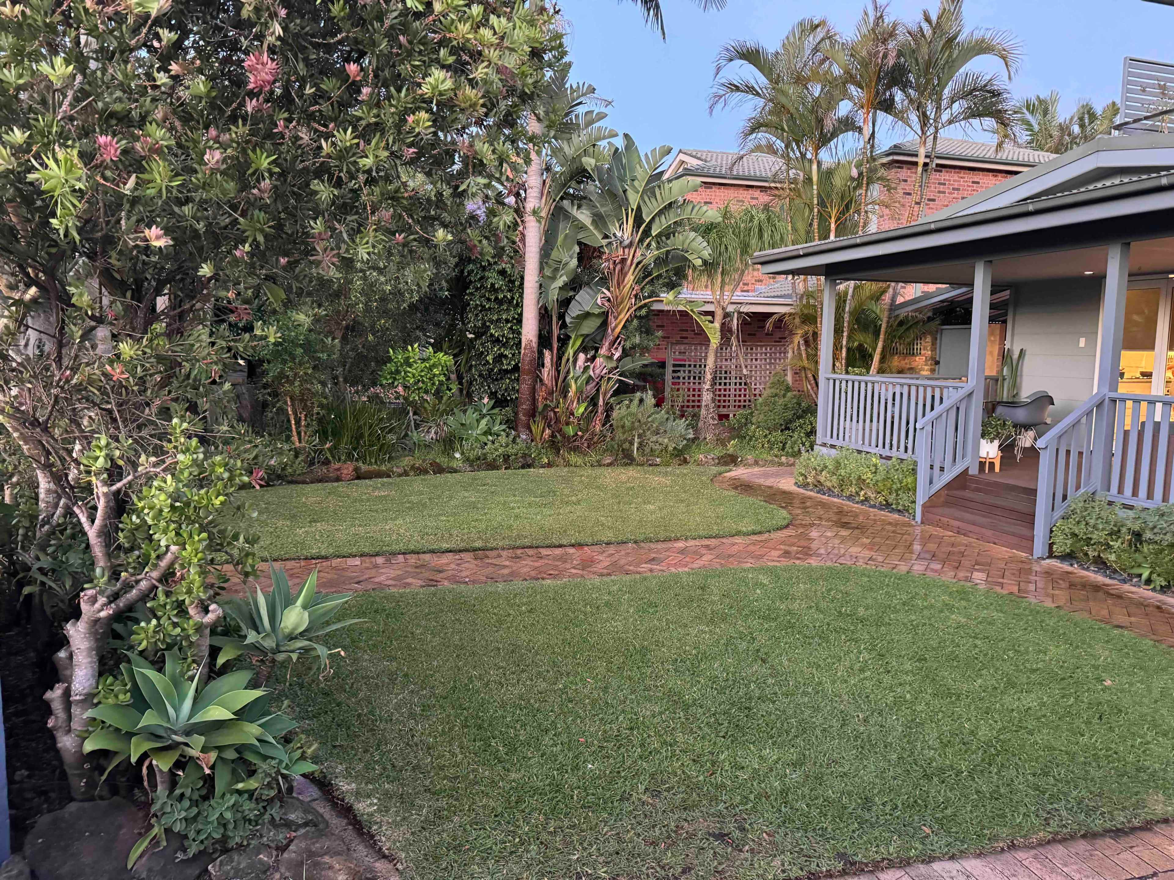 The image shows a well-maintained garden featuring a neatly mowed lawn, tropical plants, and a porch with a wooden railing.