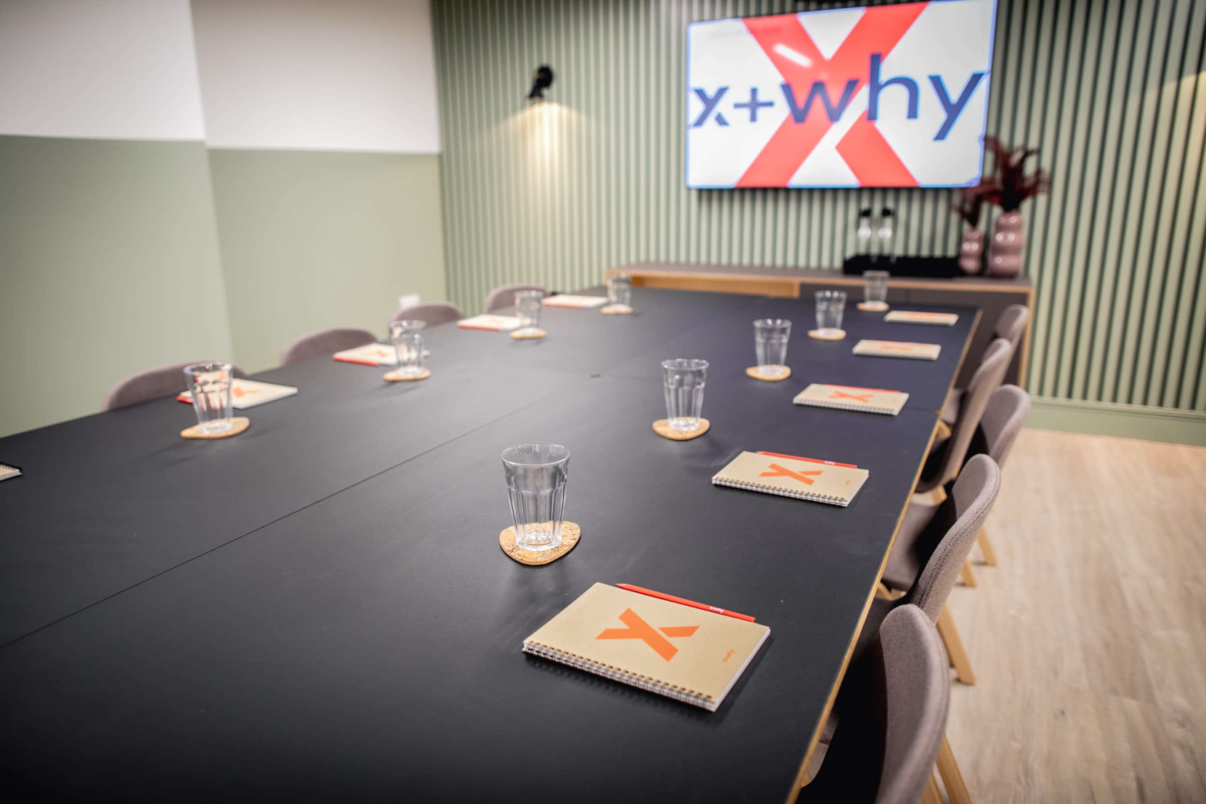 A modern meeting room features a long black table set with notebooks and glasses, accompanied by a wall-mounted screen displaying the logo "x + why."