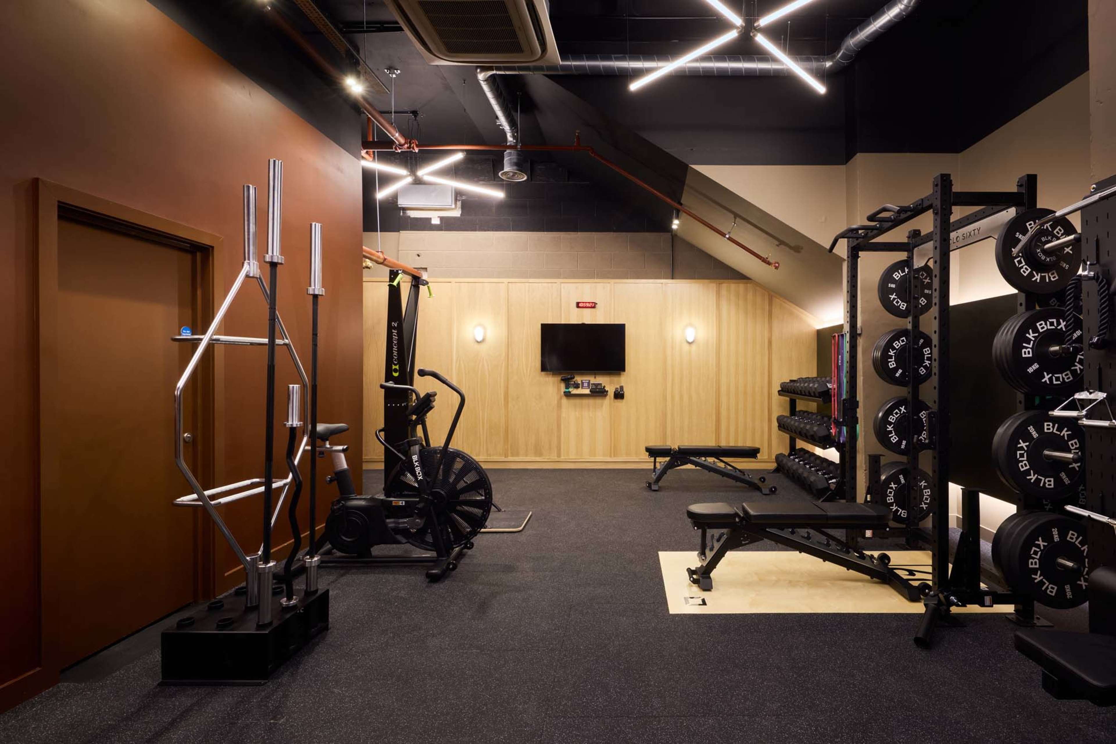 The image shows a modern gym space featuring various exercise equipment, including weight racks, a bench, and a cardio machine, against a backdrop of simple walls and ceiling lights.