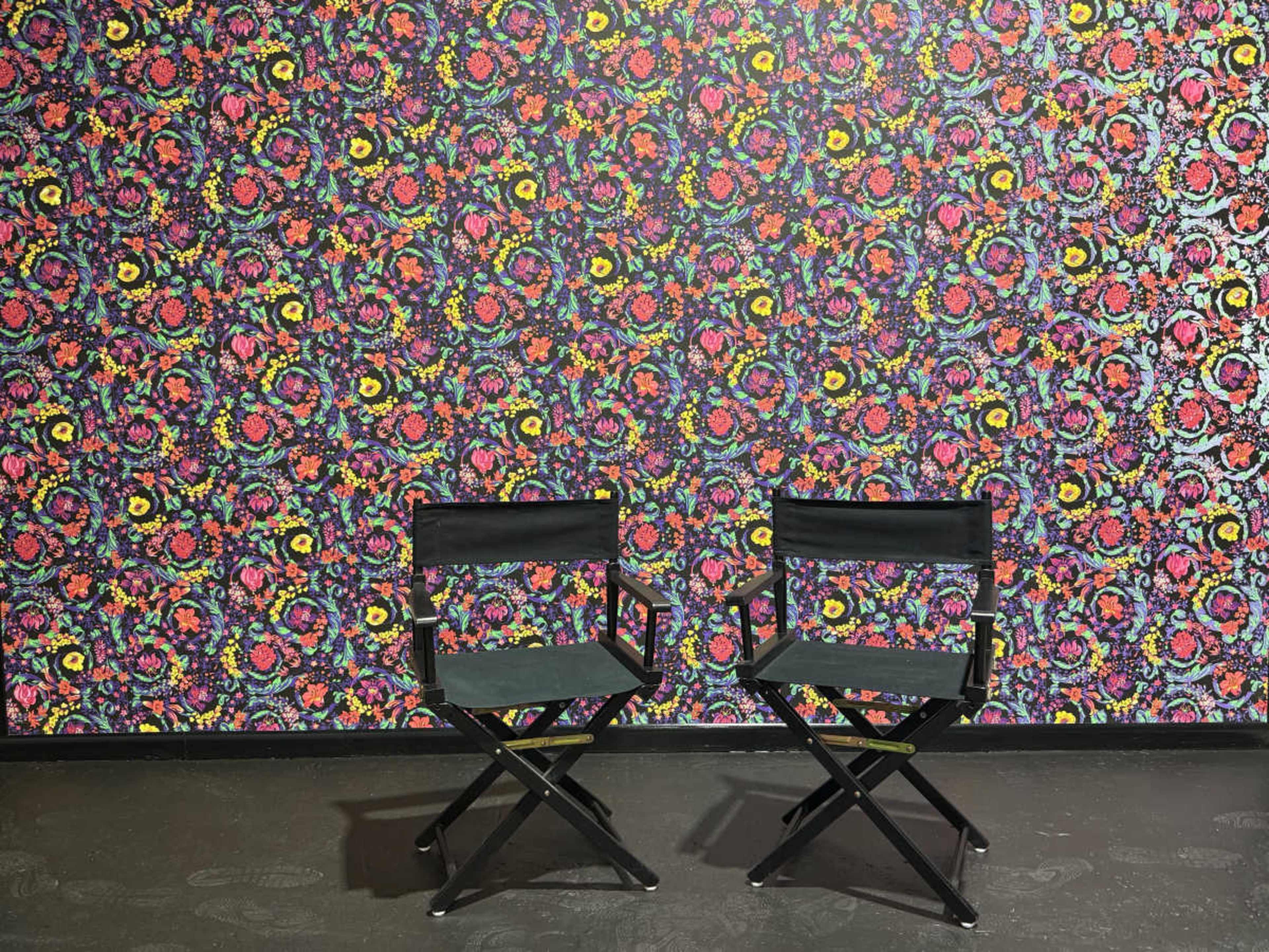 The image shows two black folding chairs positioned in front of a colorful floral-patterned wall.