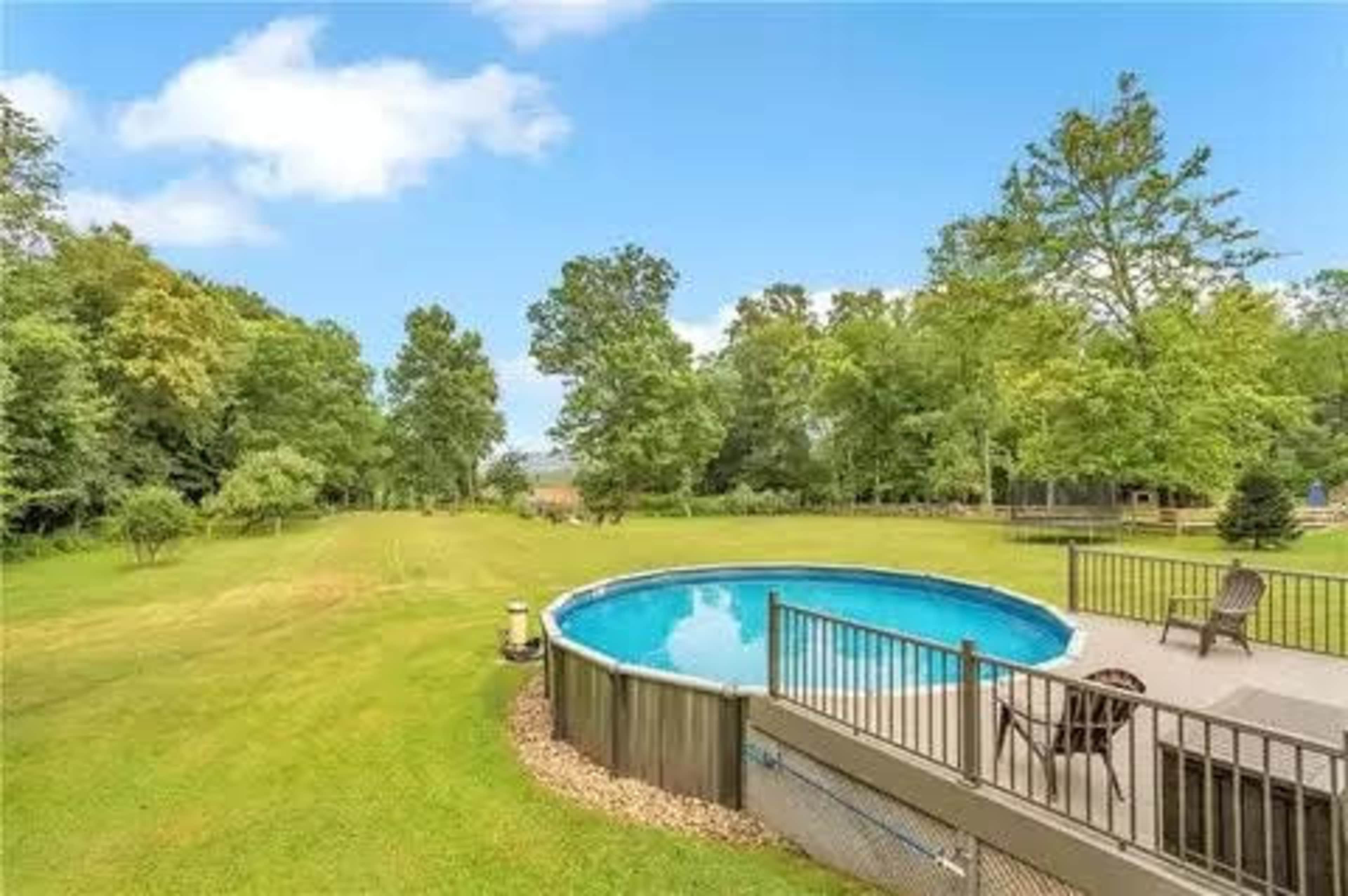 The image shows a backyard with a circular pool surrounded by a wooden deck and a lush green lawn bordered by trees.