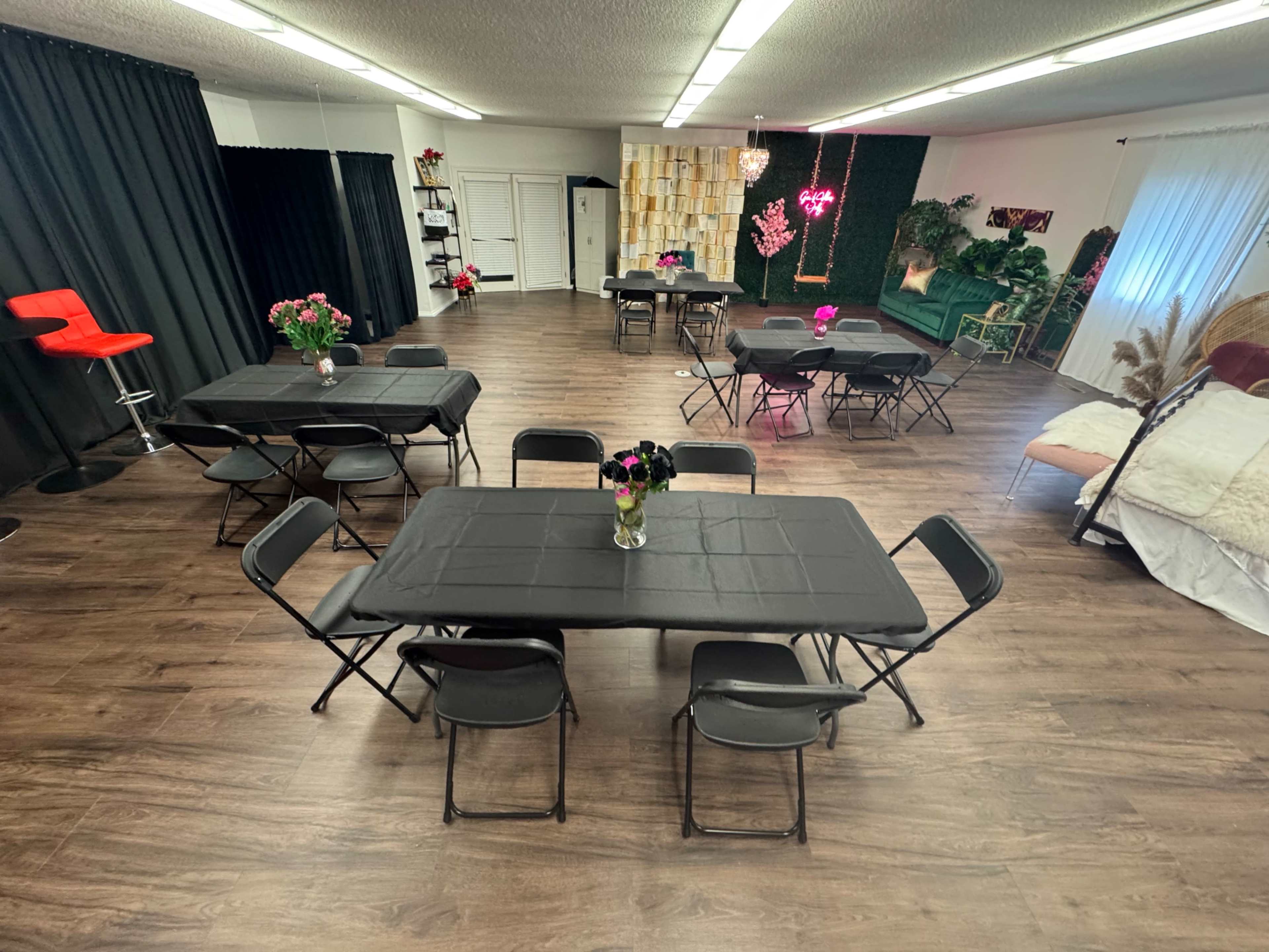The image shows an empty event space with several black tables arranged in a grid, surrounded by black curtains and decorated with potted plants and flowers.
