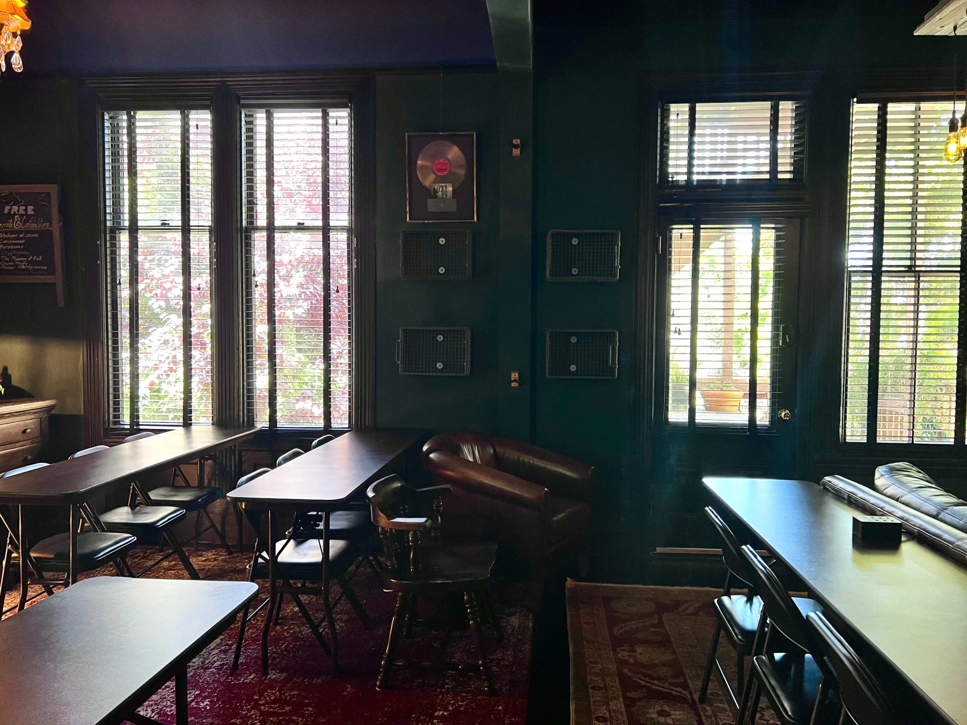 A dimly lit room with several tables and chairs arranged, large windows with blinds, and a leather couch against the wall featuring music memorabilia.