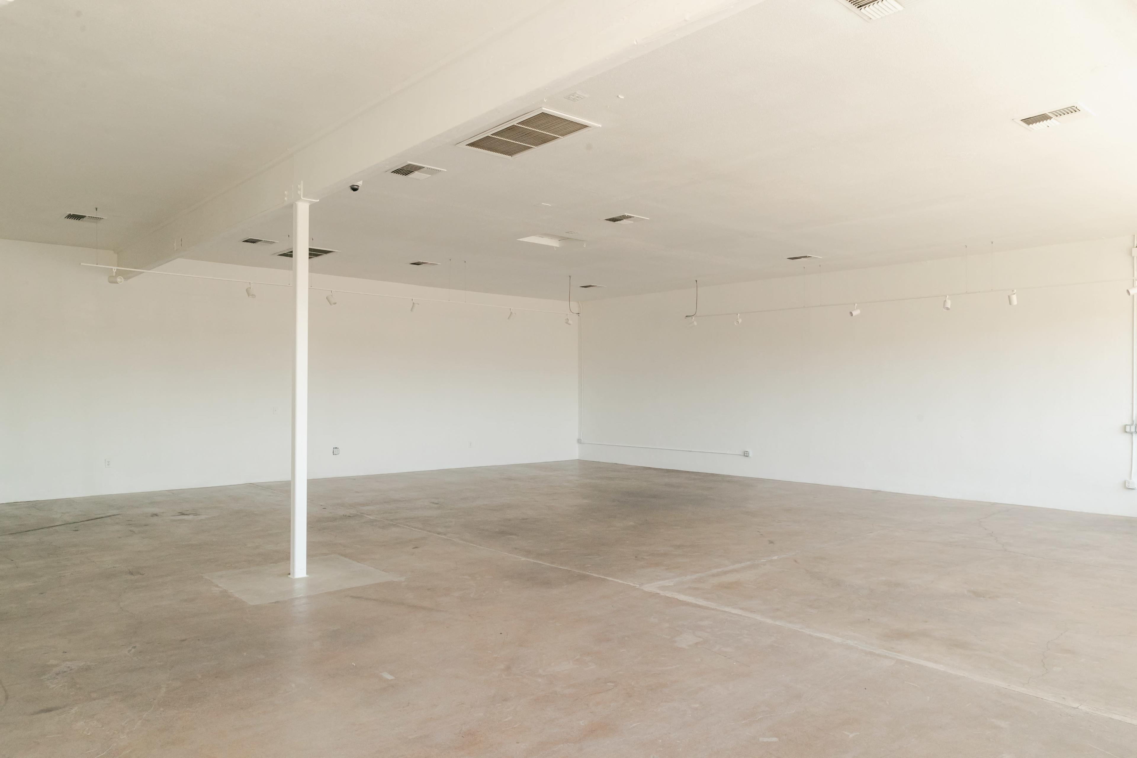 The image shows an empty room with concrete flooring and white walls, featuring a central support column and overhead lighting.