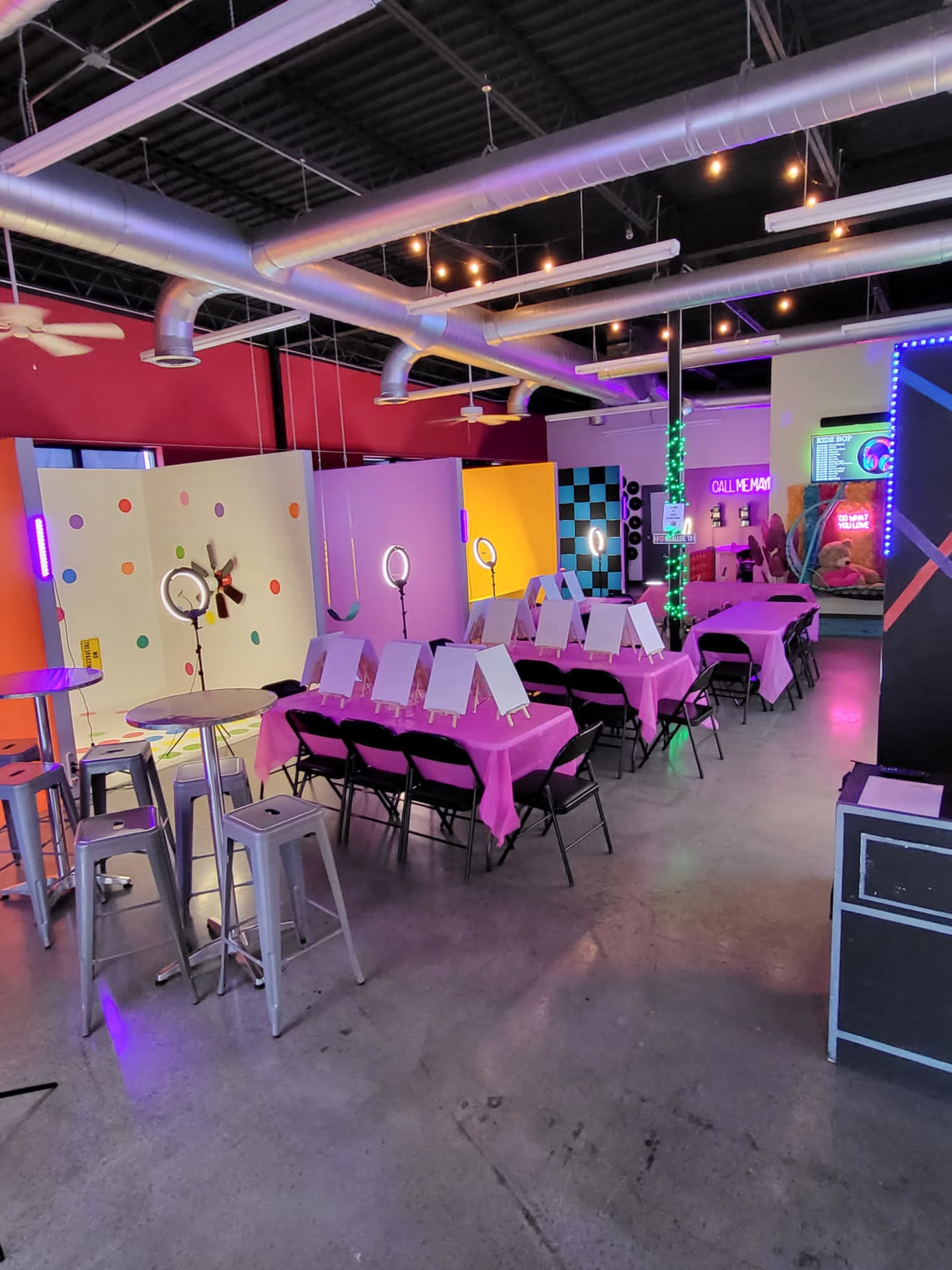 The image shows a brightly colored event space set up with tables covered in pink cloth, each featuring an easel, surrounded by colorful decorations and neon lights.