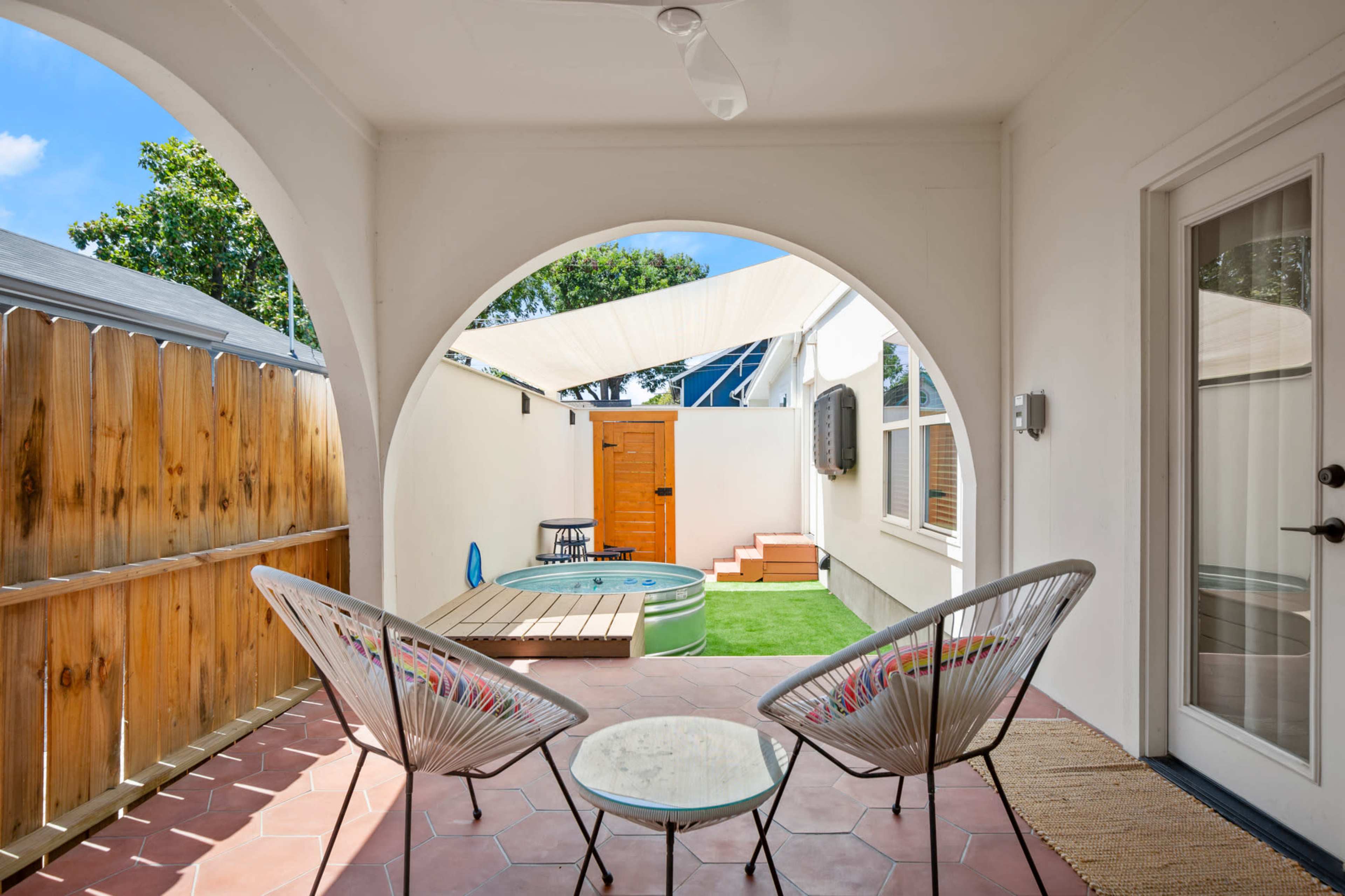 A simple outdoor patio features two wicker chairs with a glass table, surrounded by a wooden fence and an entrance to a small lawn area with steps leading up to a door.