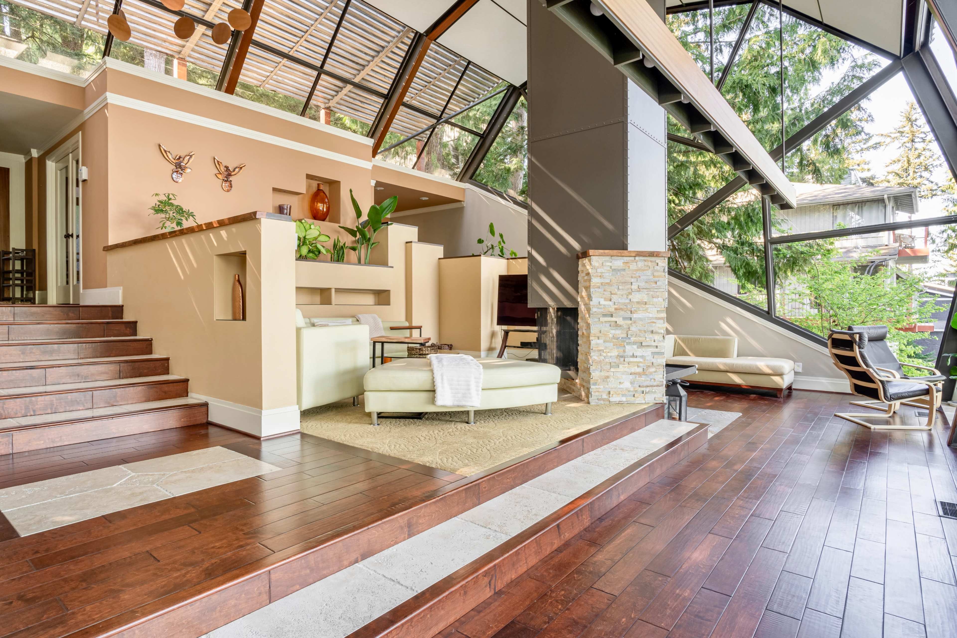 A modern living space with large glass windows, wooden flooring, and a minimalist design featuring a sectional sofa and a stone fireplace.