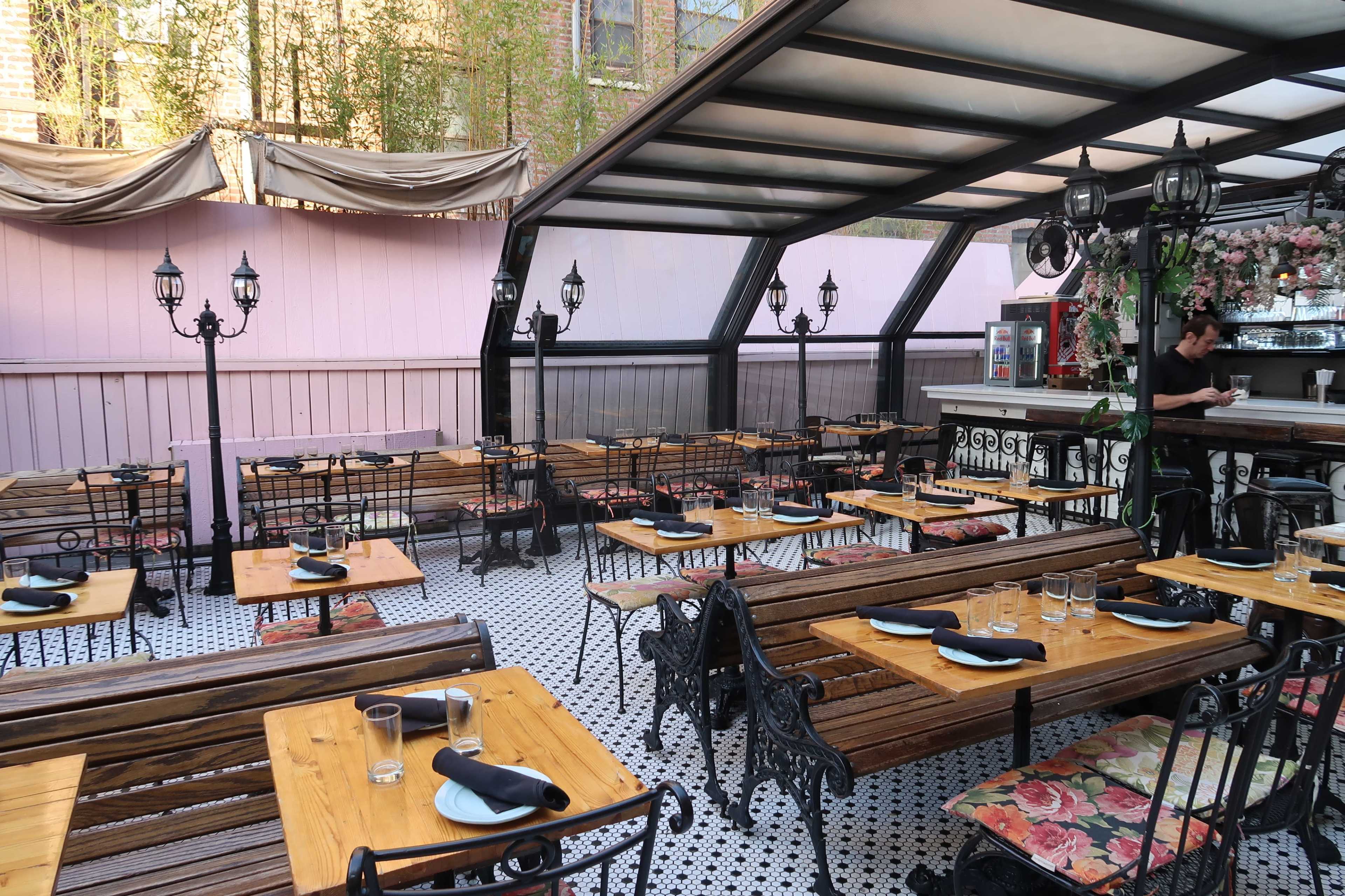 A spacious outdoor dining area with wooden tables and chairs, decorative iron lamps, and a bar in the background.