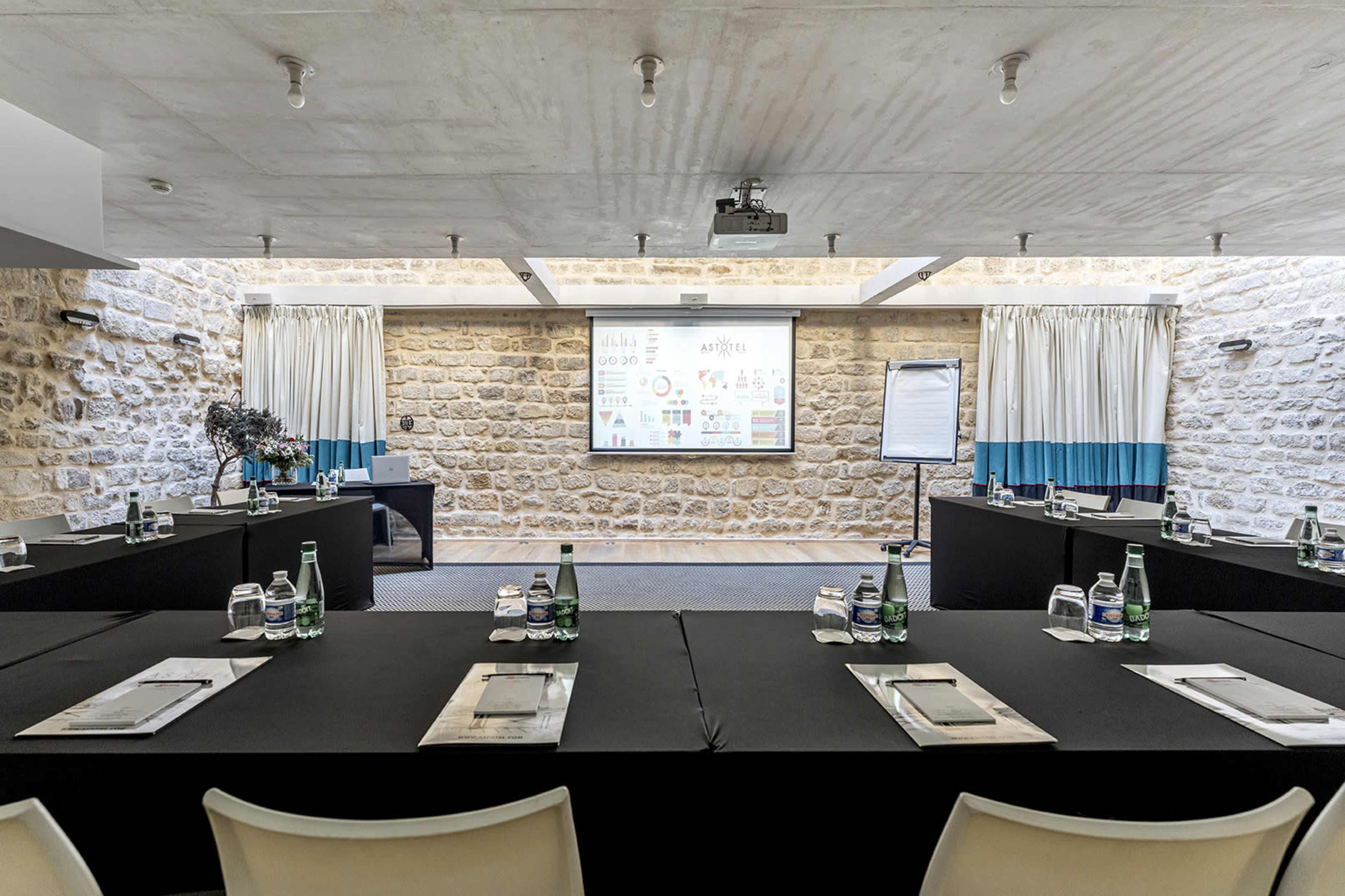 A conference room features a projector screen displaying various charts, with black-covered tables arranged in a U-shape and equipped with water bottles and notepads.