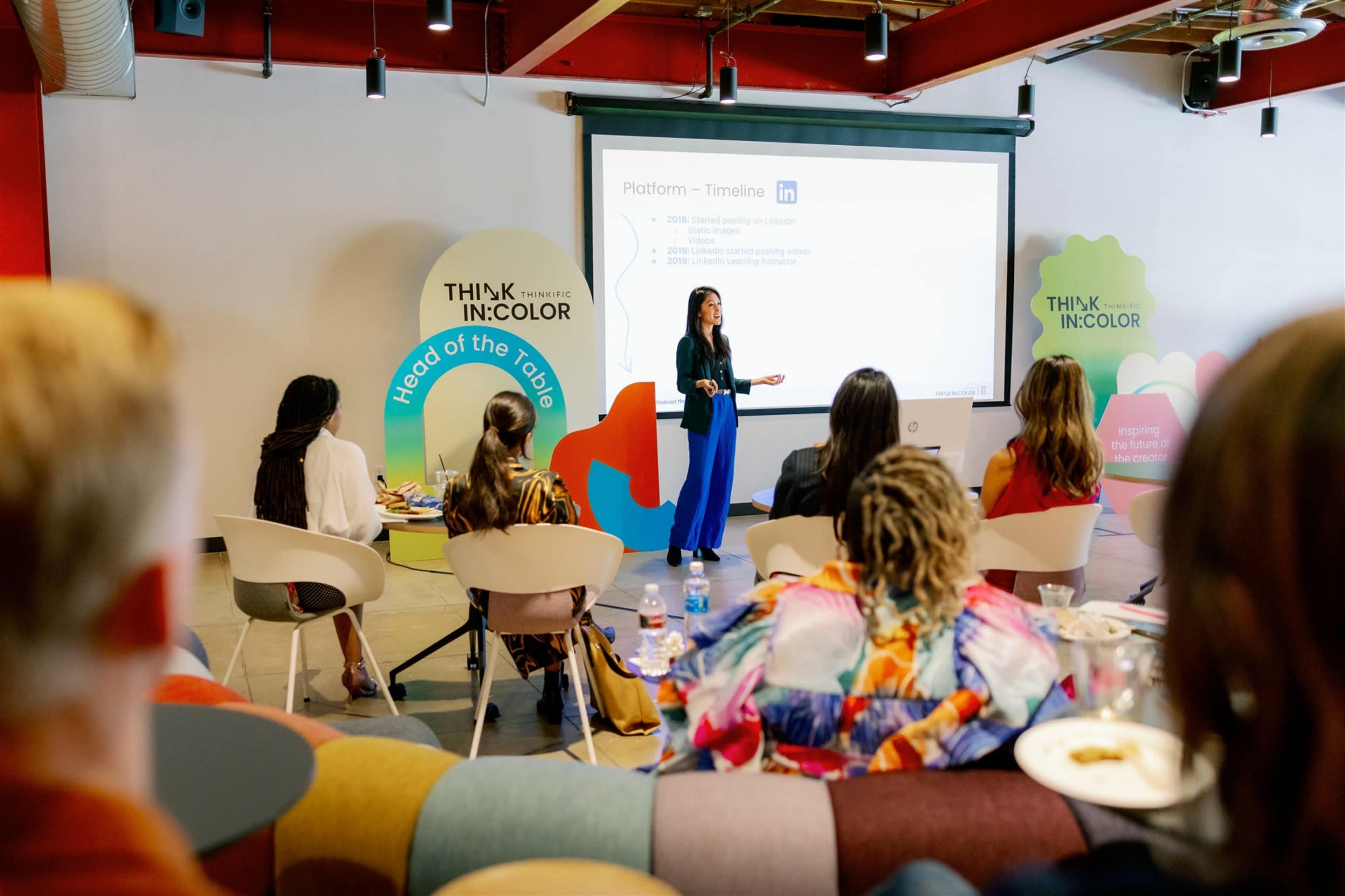 A woman presents information on a screen to a seated audience in a modern meeting space.
