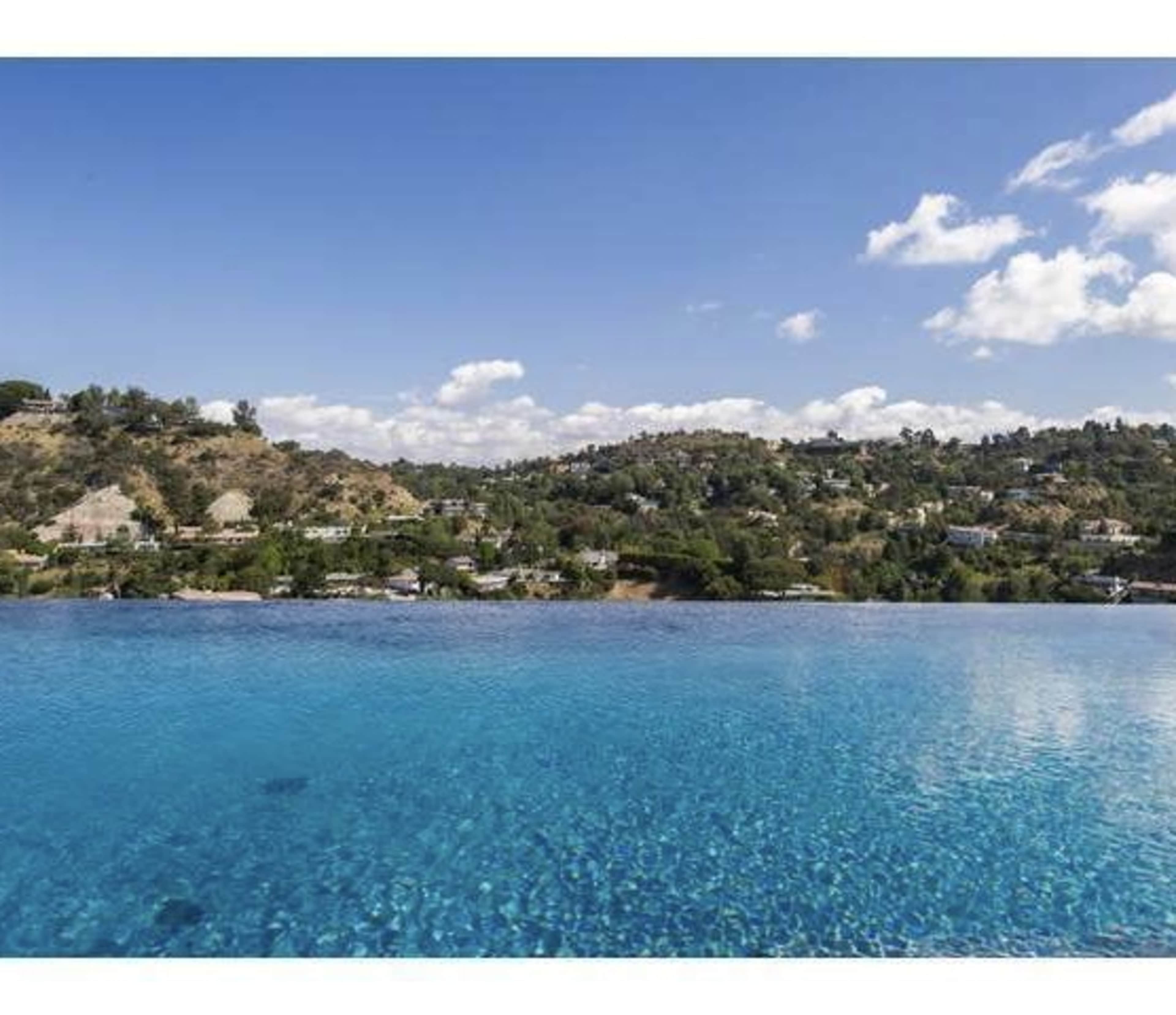 The image shows a clear blue infinity pool overlooking a hilly landscape with scattered homes under a partly cloudy sky.