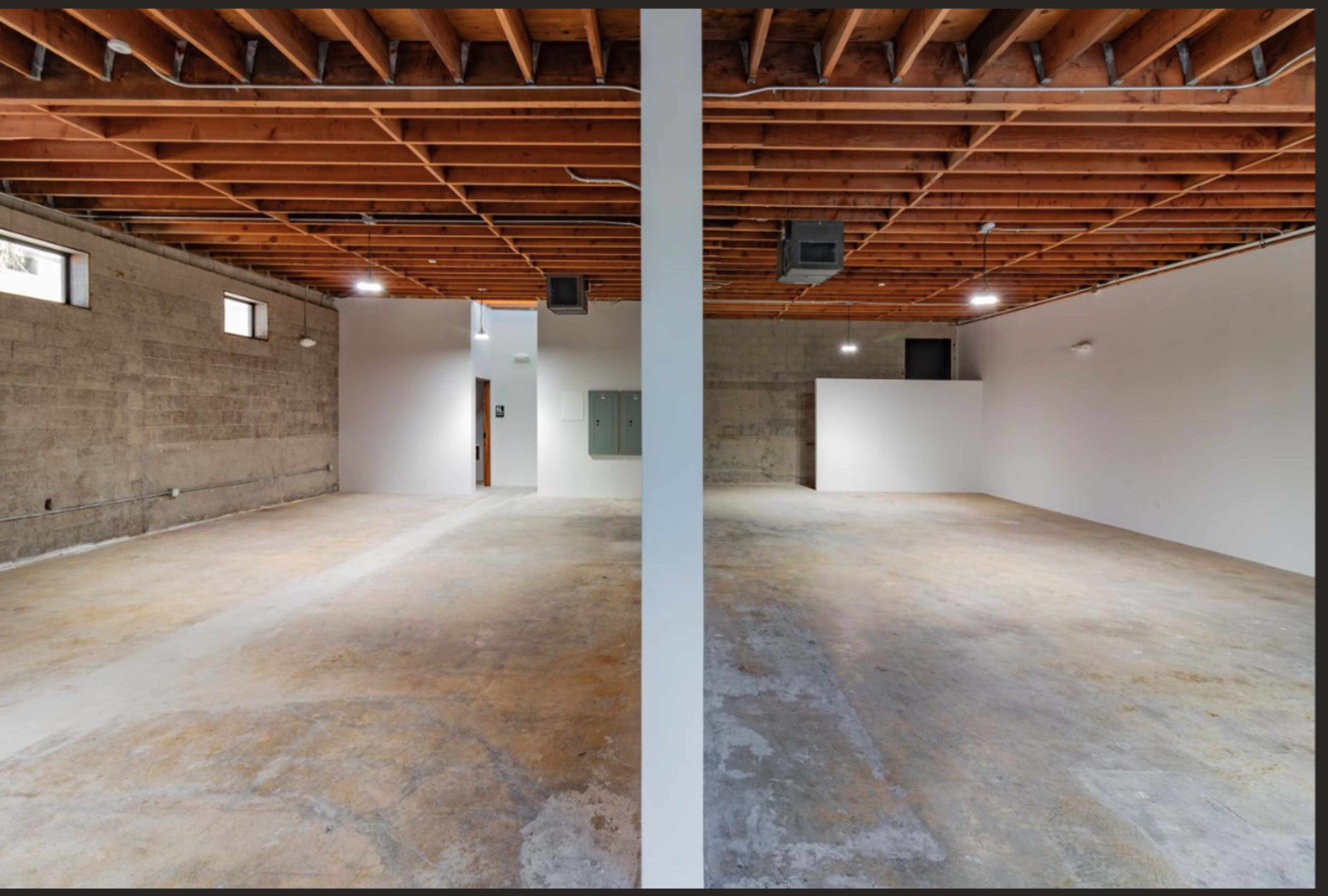 The image shows a large, empty interior space divided by a central pillar, with exposed wooden beams above and concrete walls.