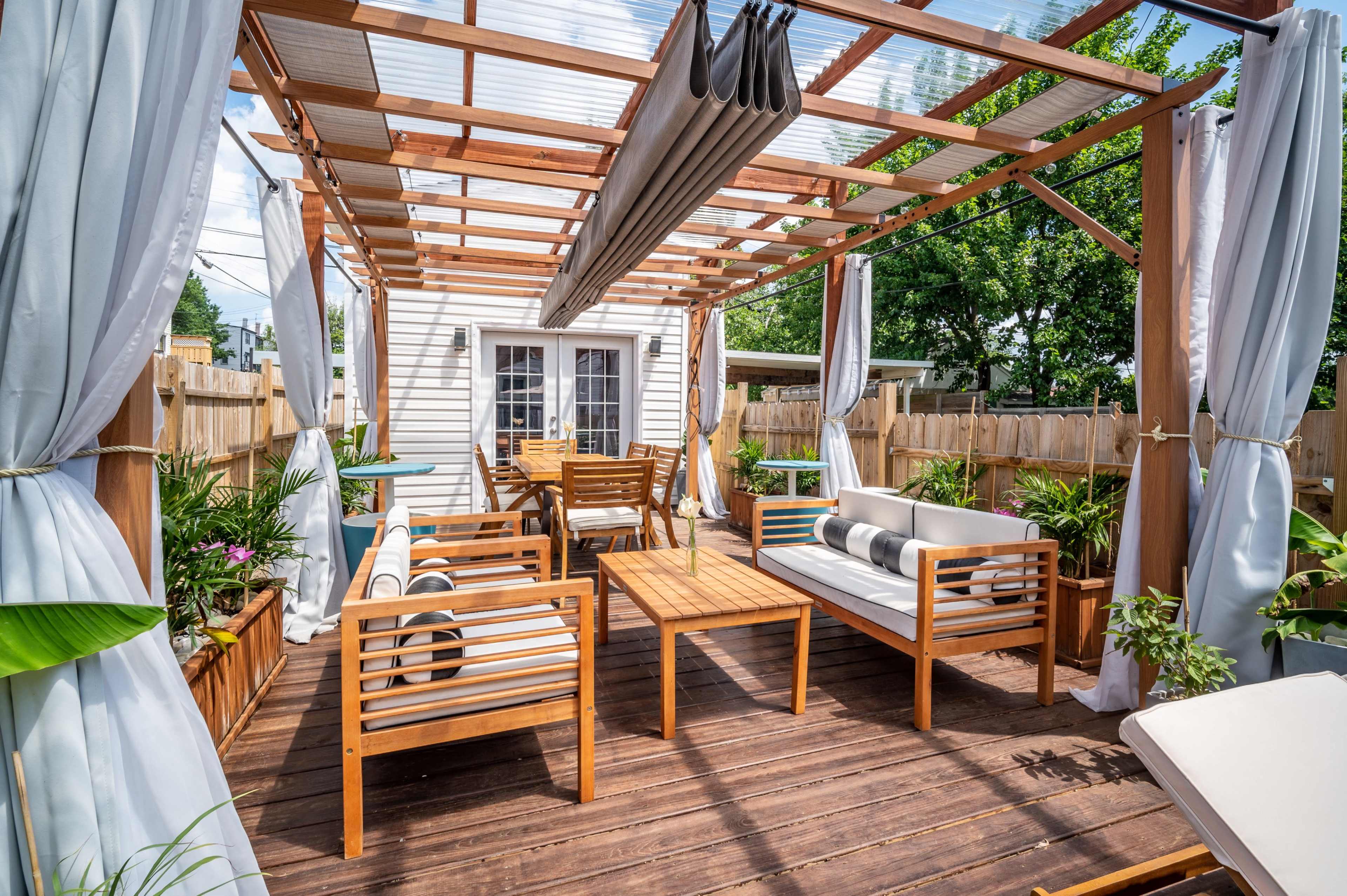 A wooden deck patio area with a pergola, featuring seating arrangements, a dining table, and potted plants.