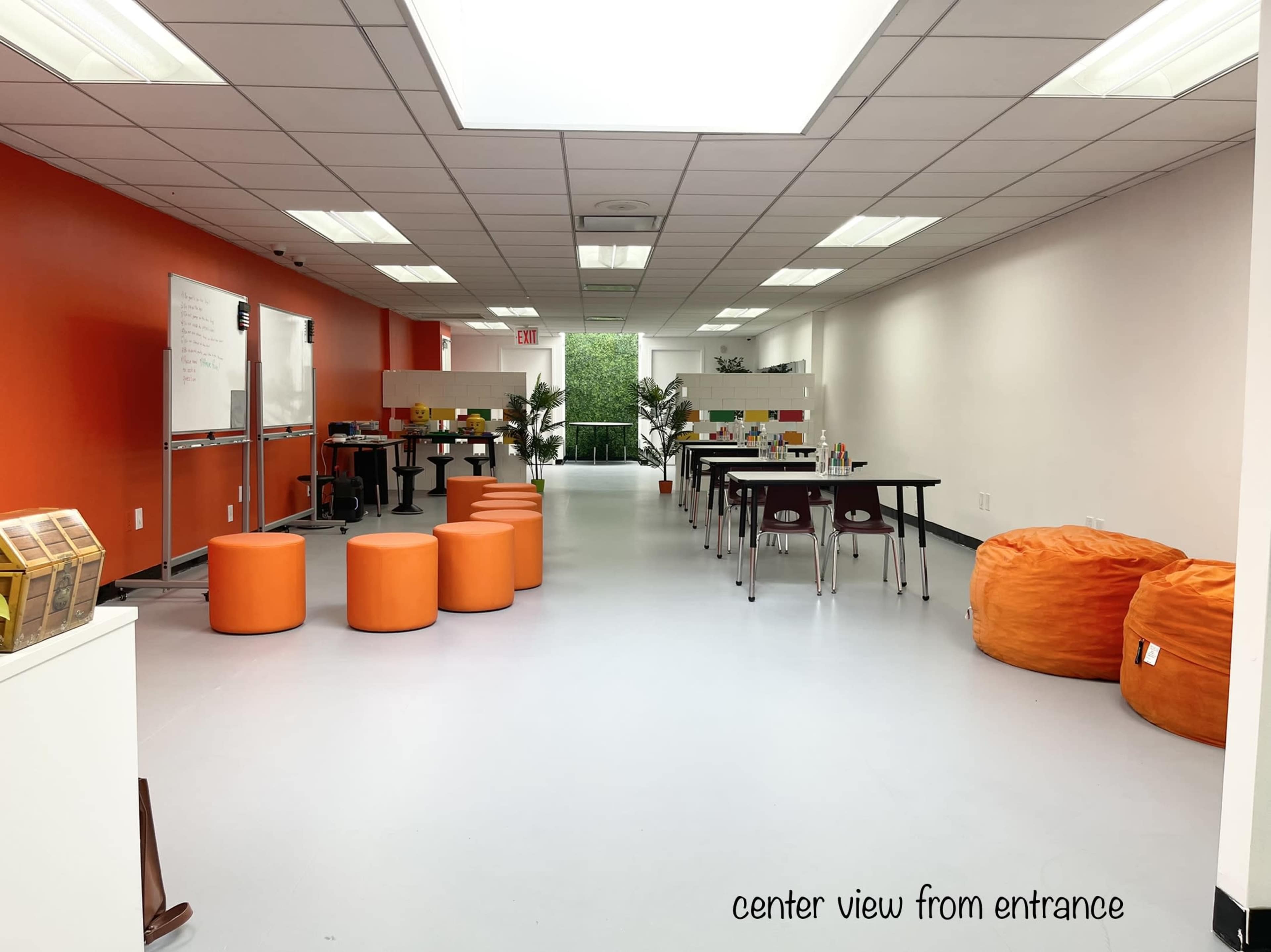 A brightly lit room featuring orange poufs, tables, and a whiteboard along the walls, with greenery visible through a large window at the far end.