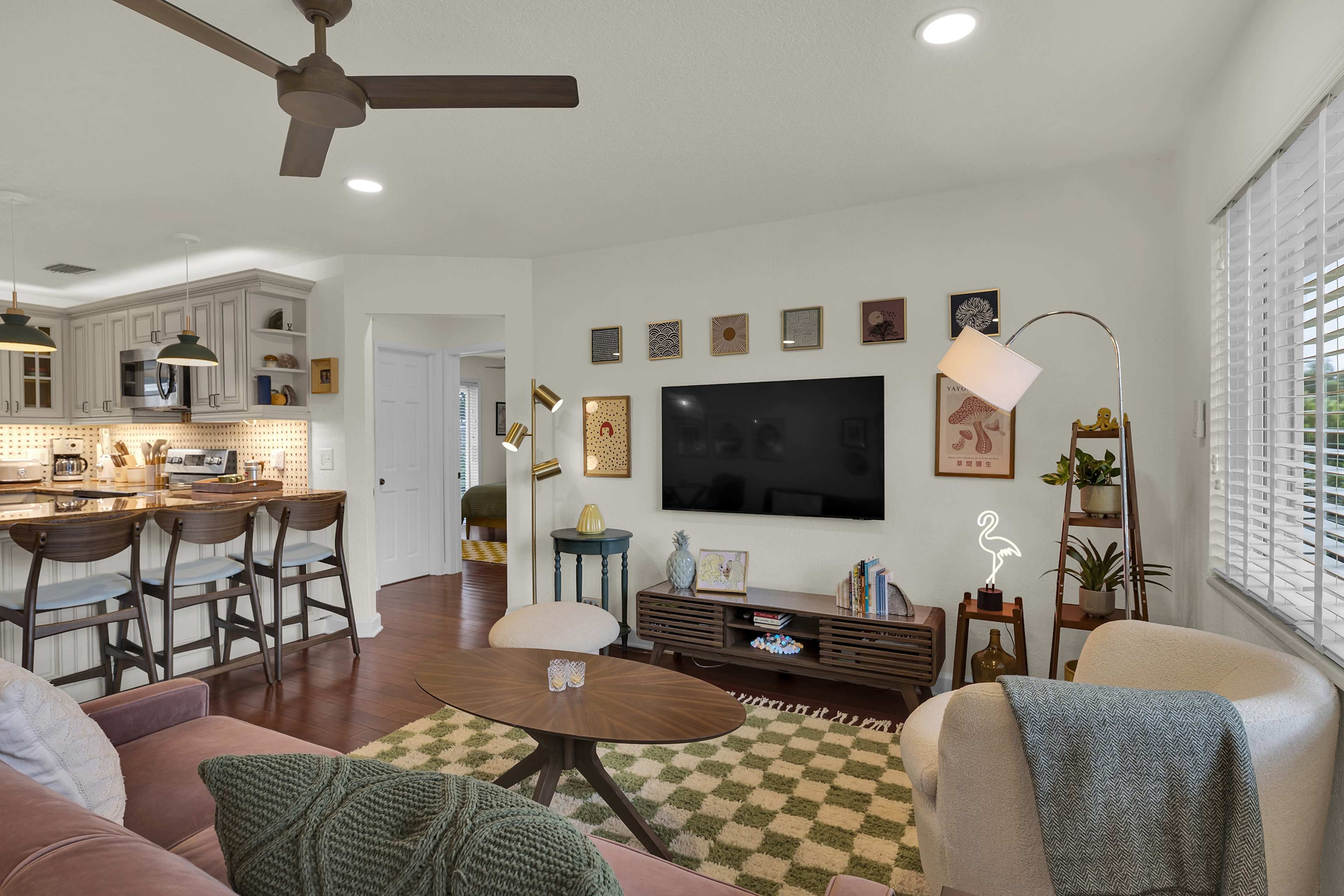 The image shows a modern living room with a brown sofa, a coffee table on a patterned rug, and a mounted television on the wall, adjacent to a kitchen area with bar seating.