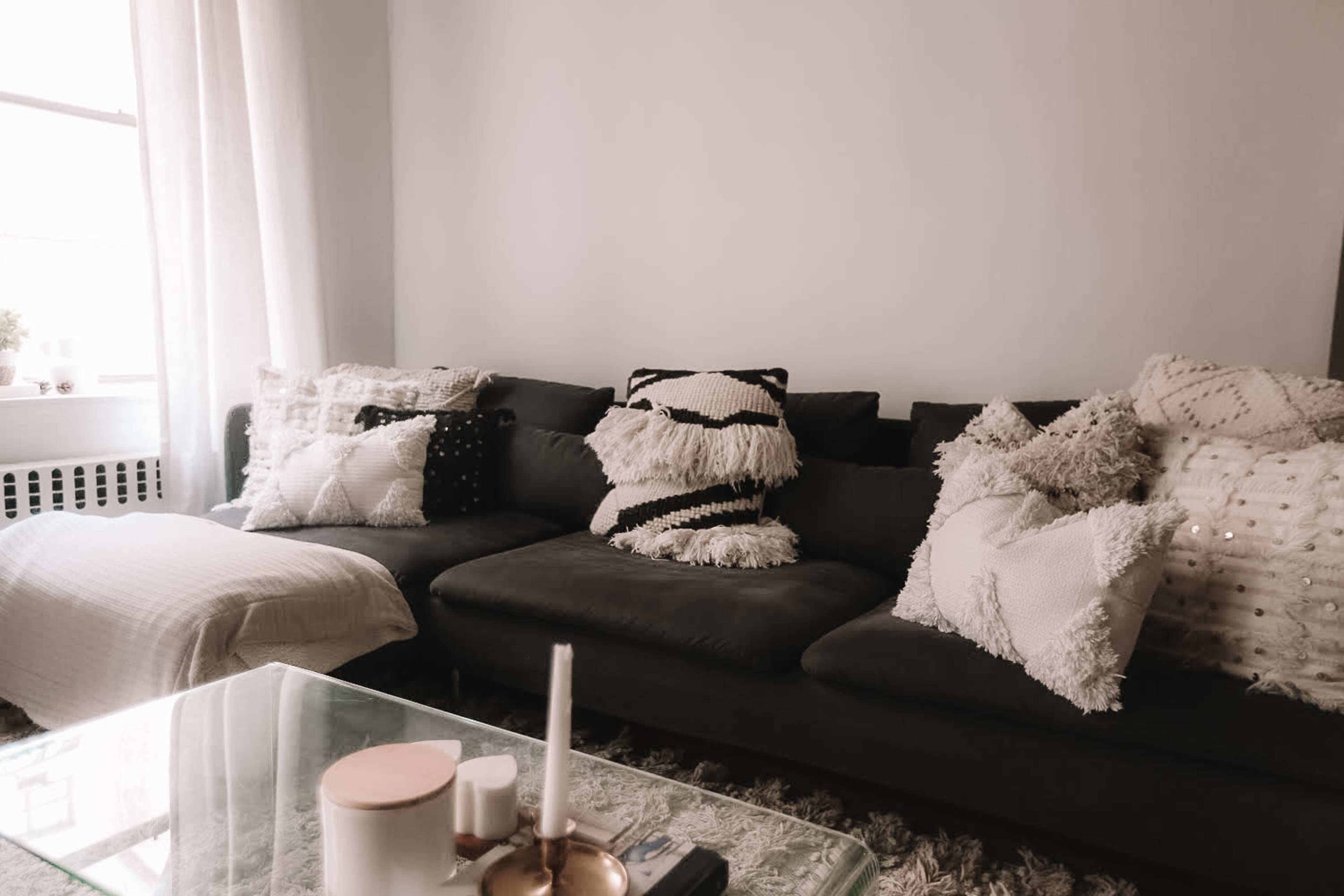 A cozy living room features a dark sofa adorned with various textured pillows and a glass coffee table with a candle and decorative objects.