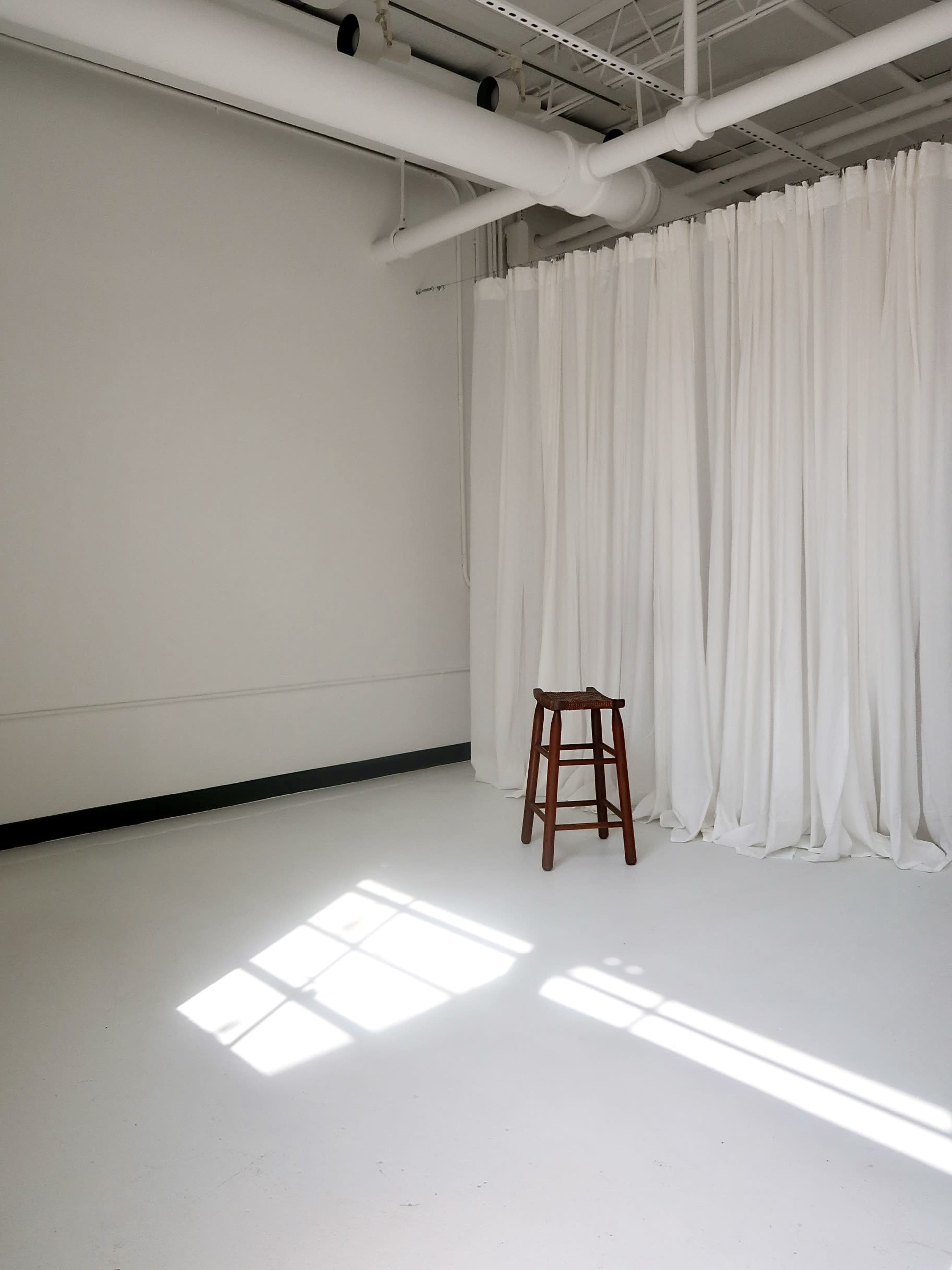 A wooden stool stands in an empty room with white walls and a white curtain backdrop.
