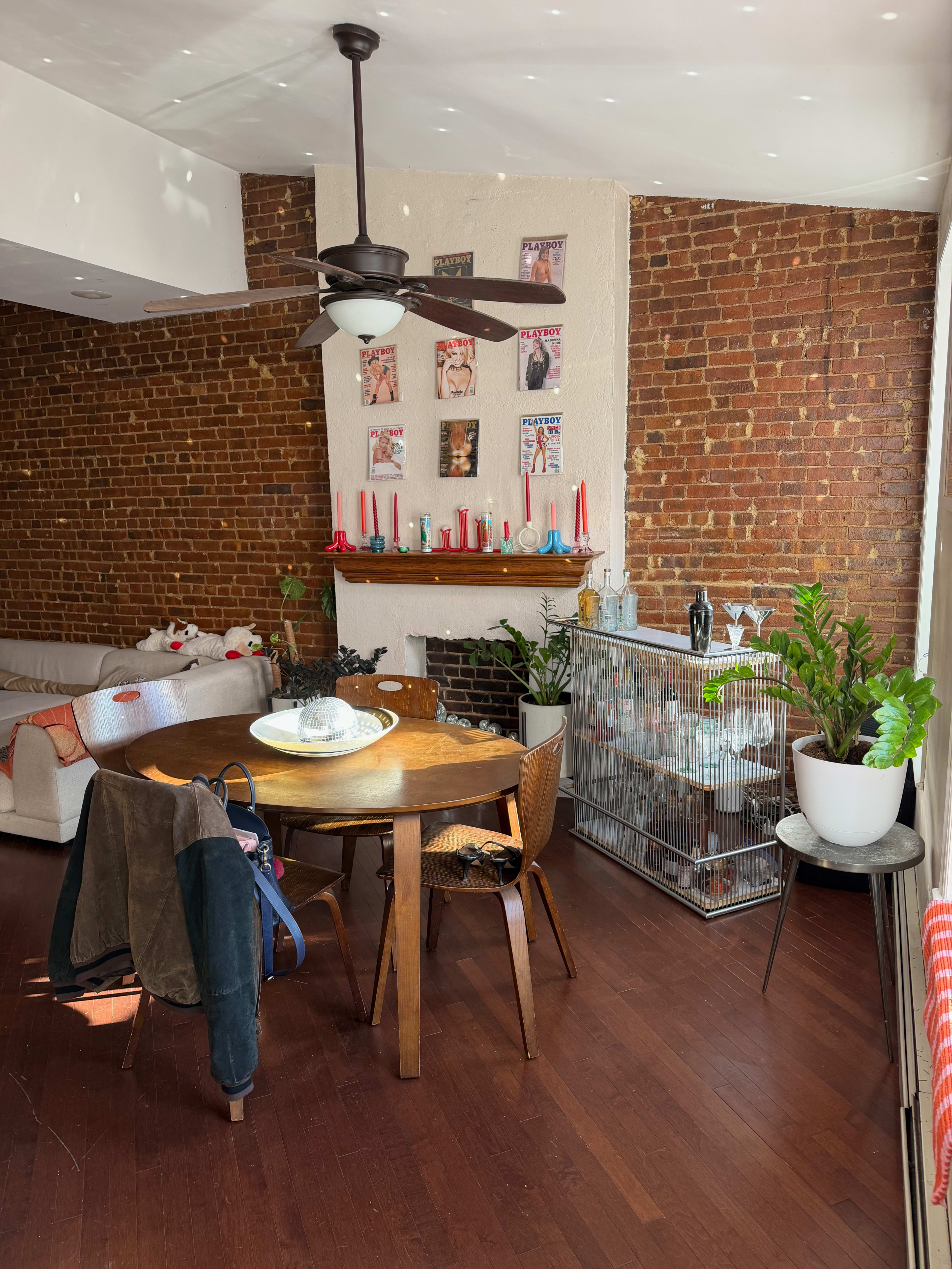 A wooden dining table with chairs is positioned in a room featuring exposed brick walls, a ceiling fan, and decorative items on a mantel.