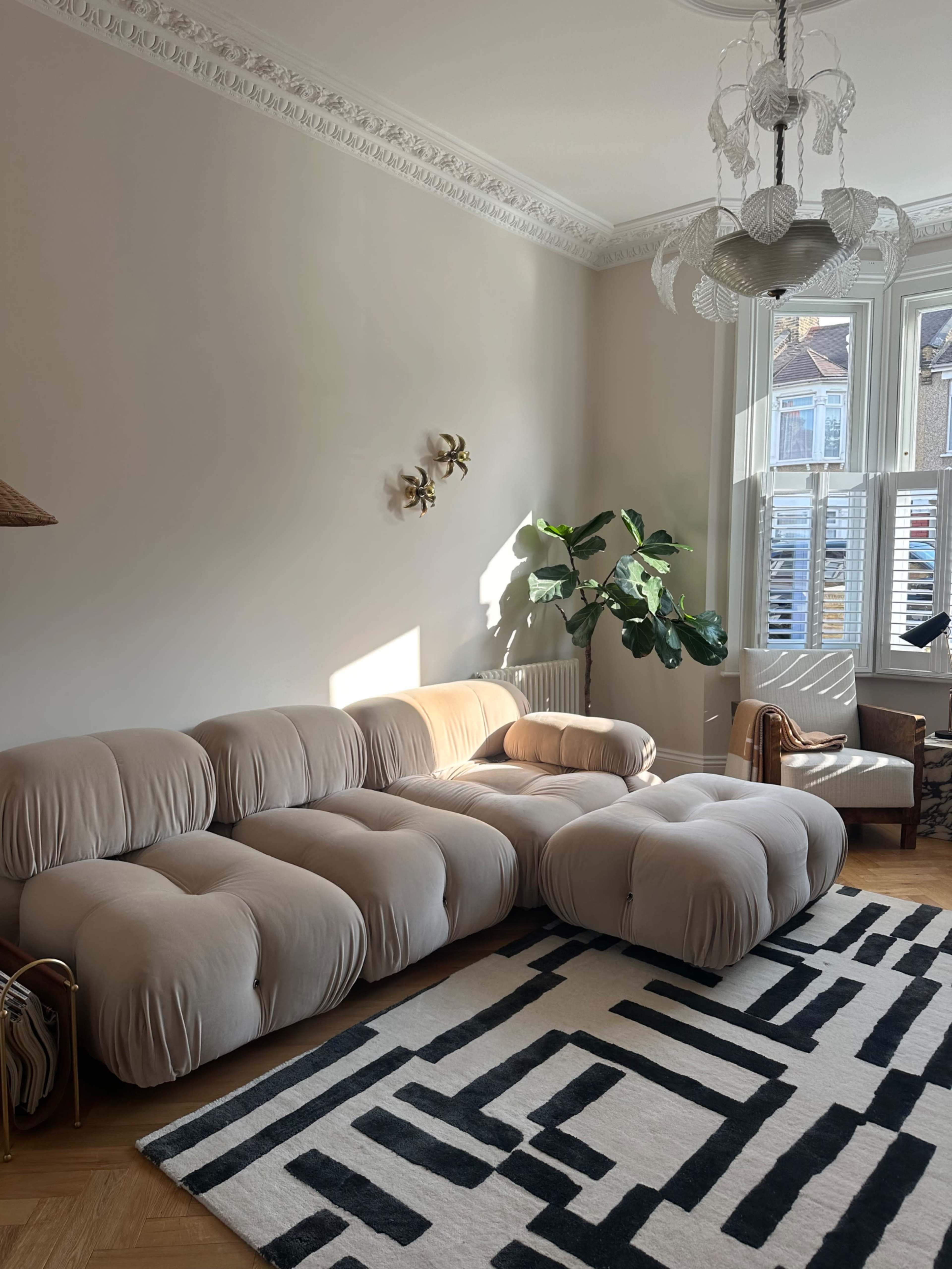 The image shows a modern living room with a large, soft beige sectional sofa, a patterned rug, a potted plant, and natural light streaming through the windows.
