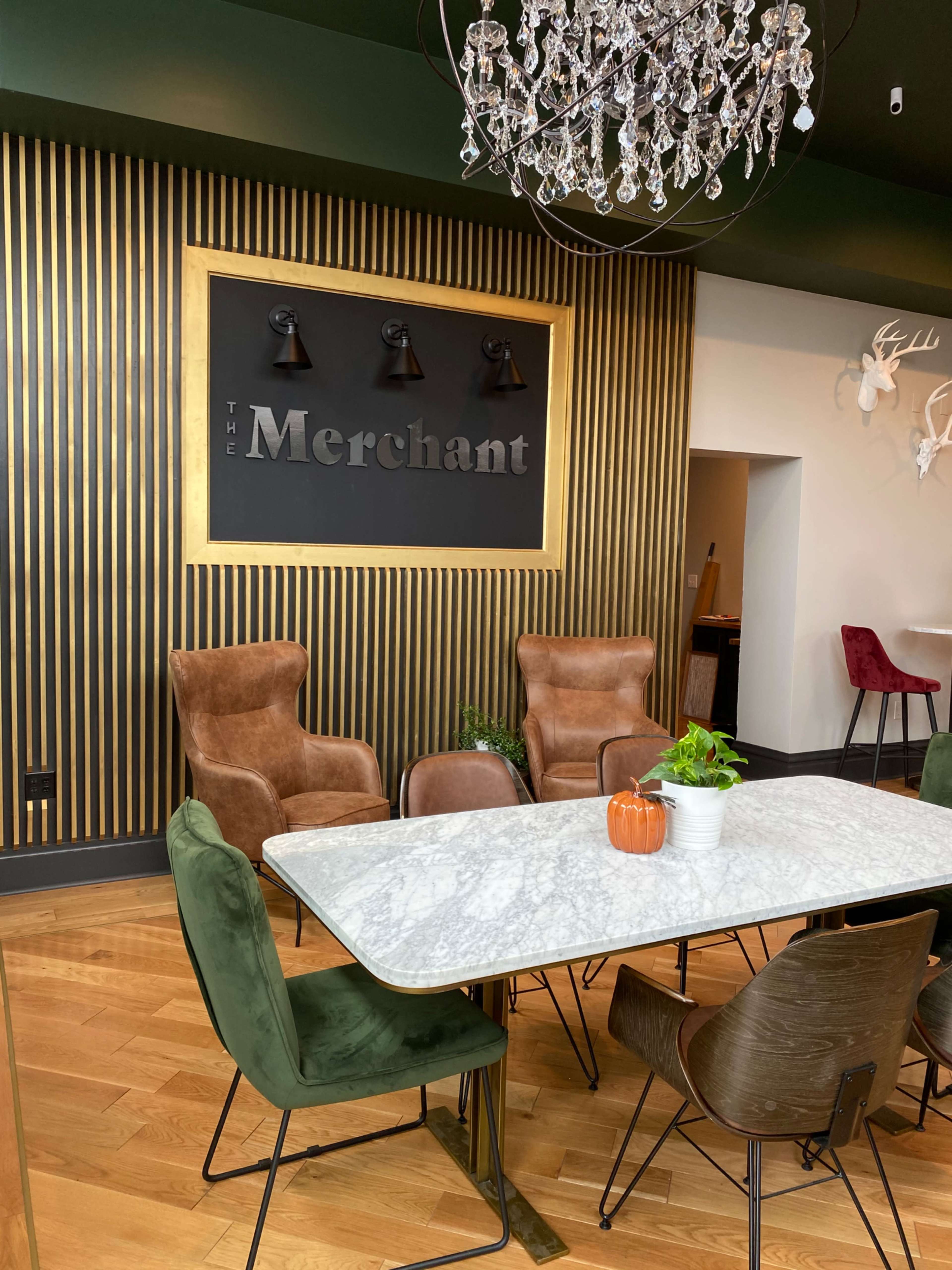 A modern dining area features a marble table surrounded by various stylish chairs, with a decorative wall displaying the name "The Merchant" and a chandelier overhead.