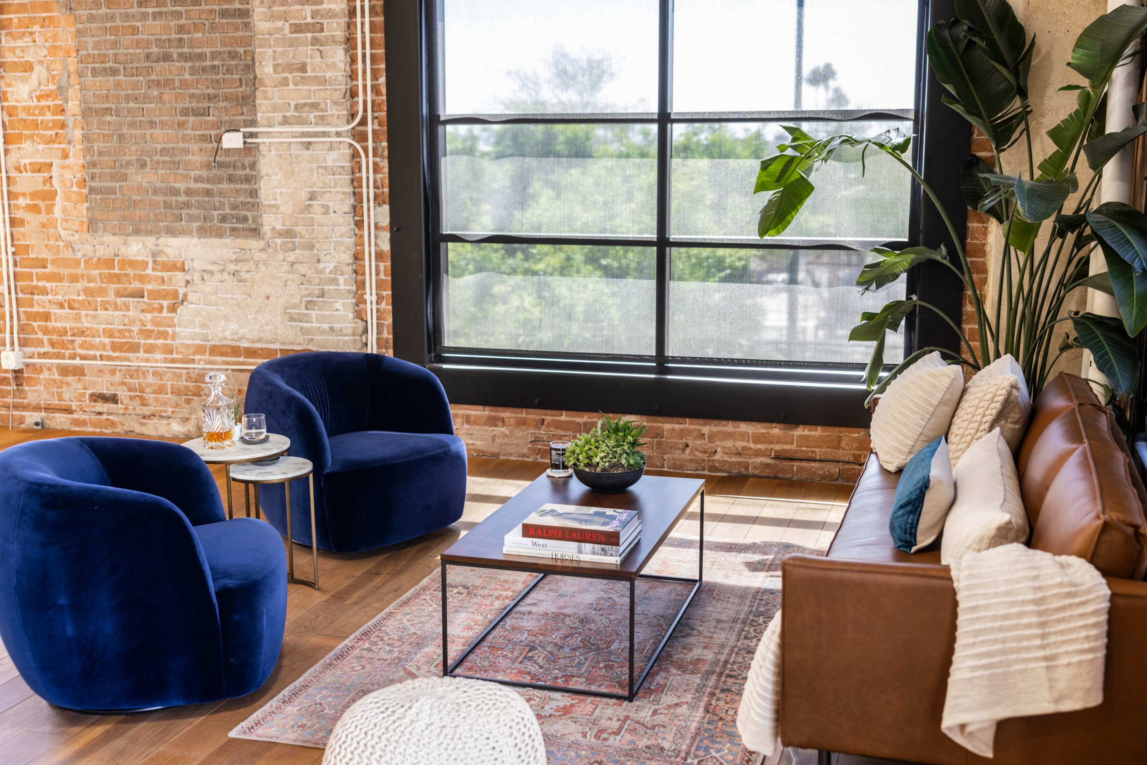 A modern living room featuring a brown leather sofa, two blue velvet chairs, a coffee table with a plant and books, and large windows allowing natural light to enter, set against a backdrop of exposed brick walls.