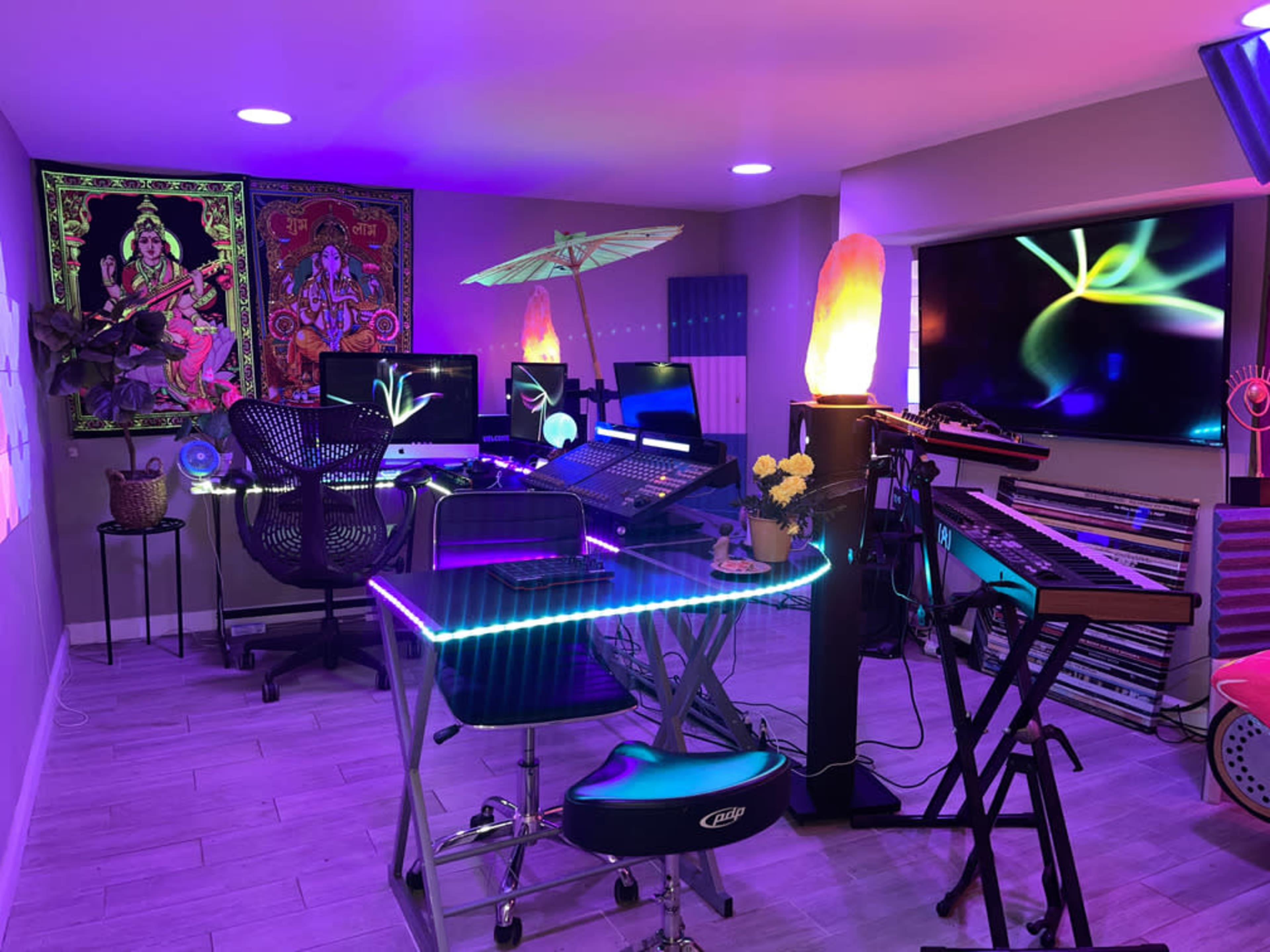 A vibrant music studio with multiple computers, keyboards, decorative lighting, and a modern glass desk.
