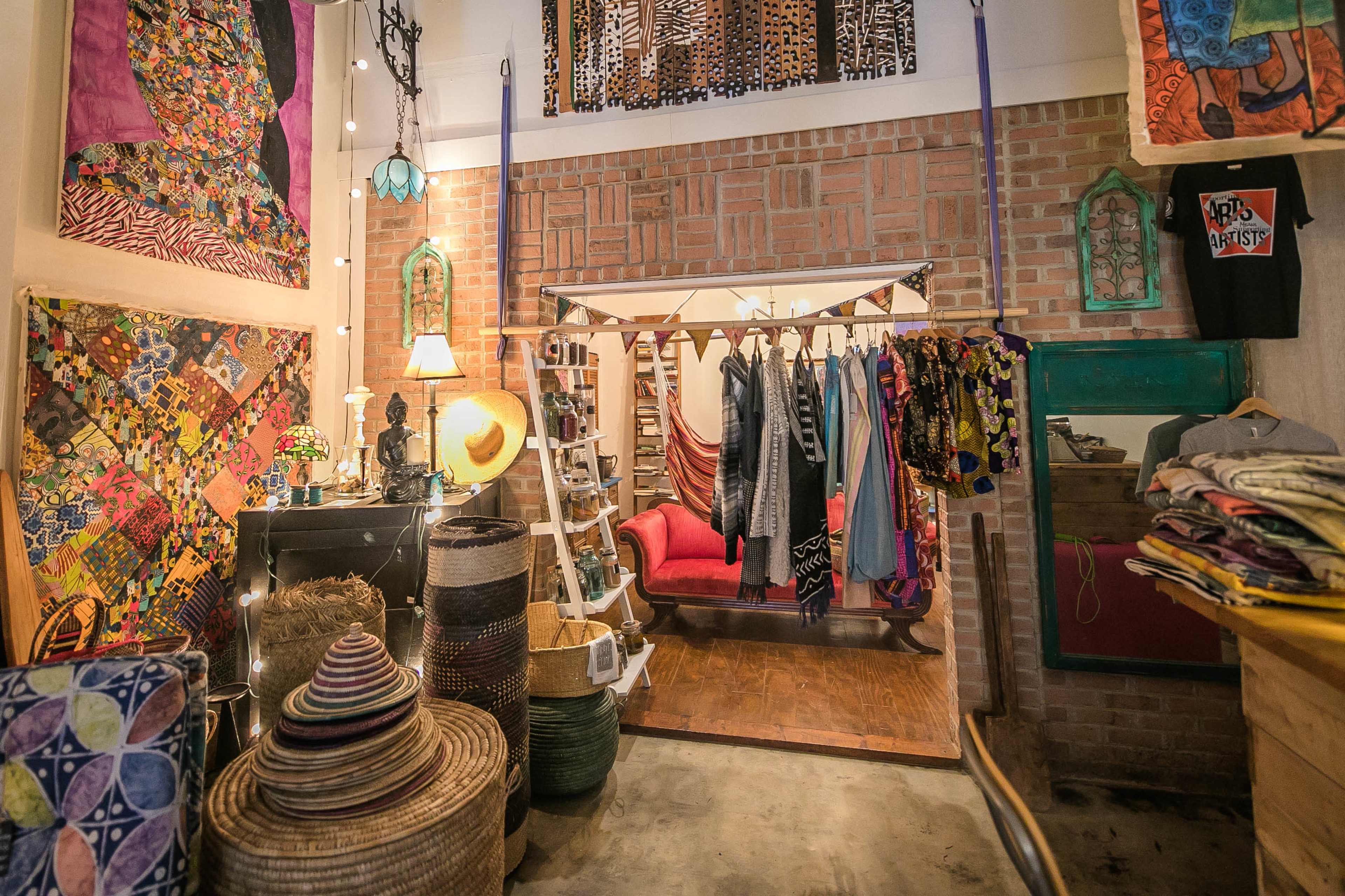 A cozy boutique displaying colorful textiles and clothing, with a brick wall backdrop and various woven baskets arranged around the room.