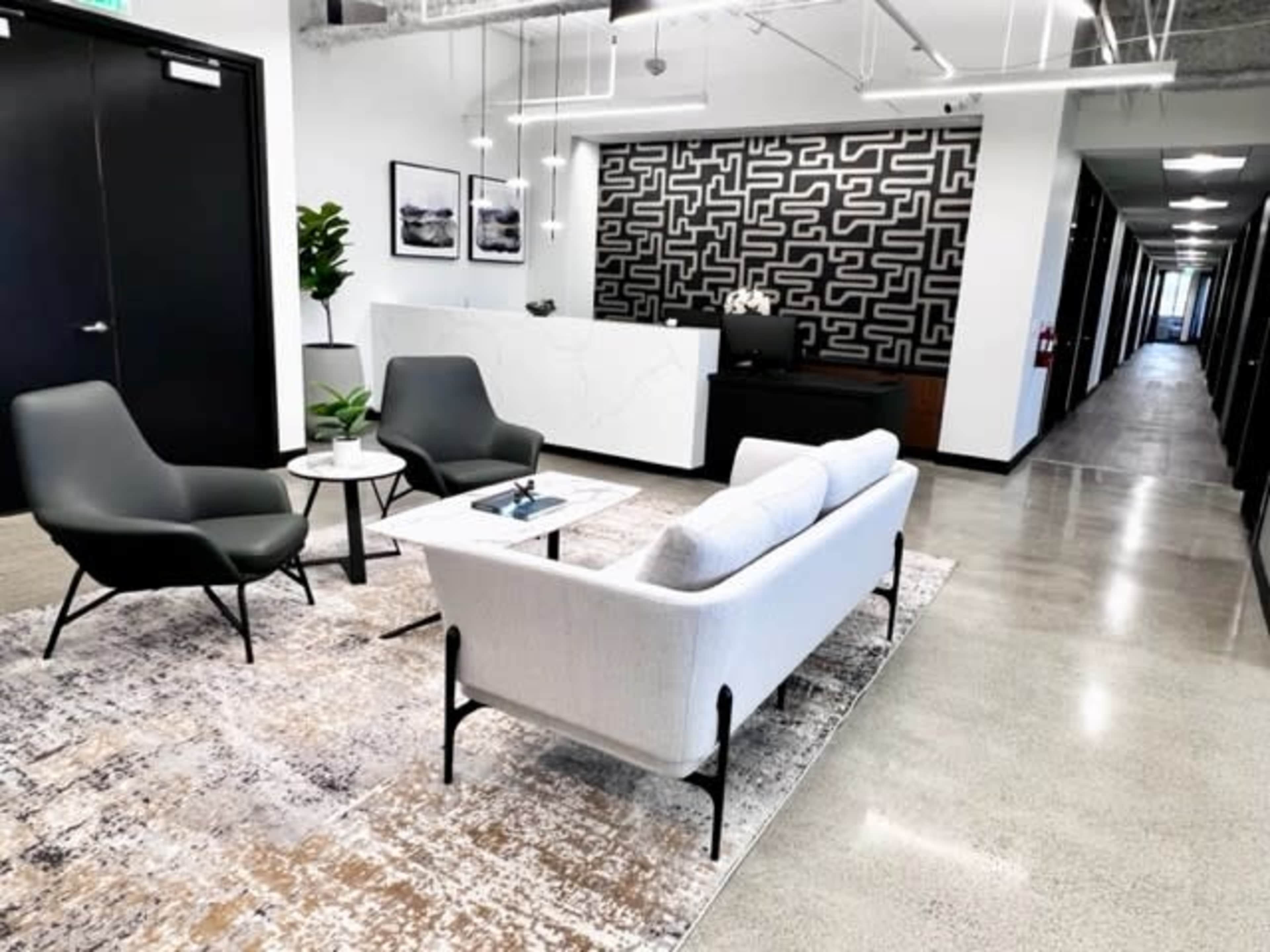 A modern office reception area with two chairs and a sofa arranged on a rug, a decorative wall, and a long hallway in the background.