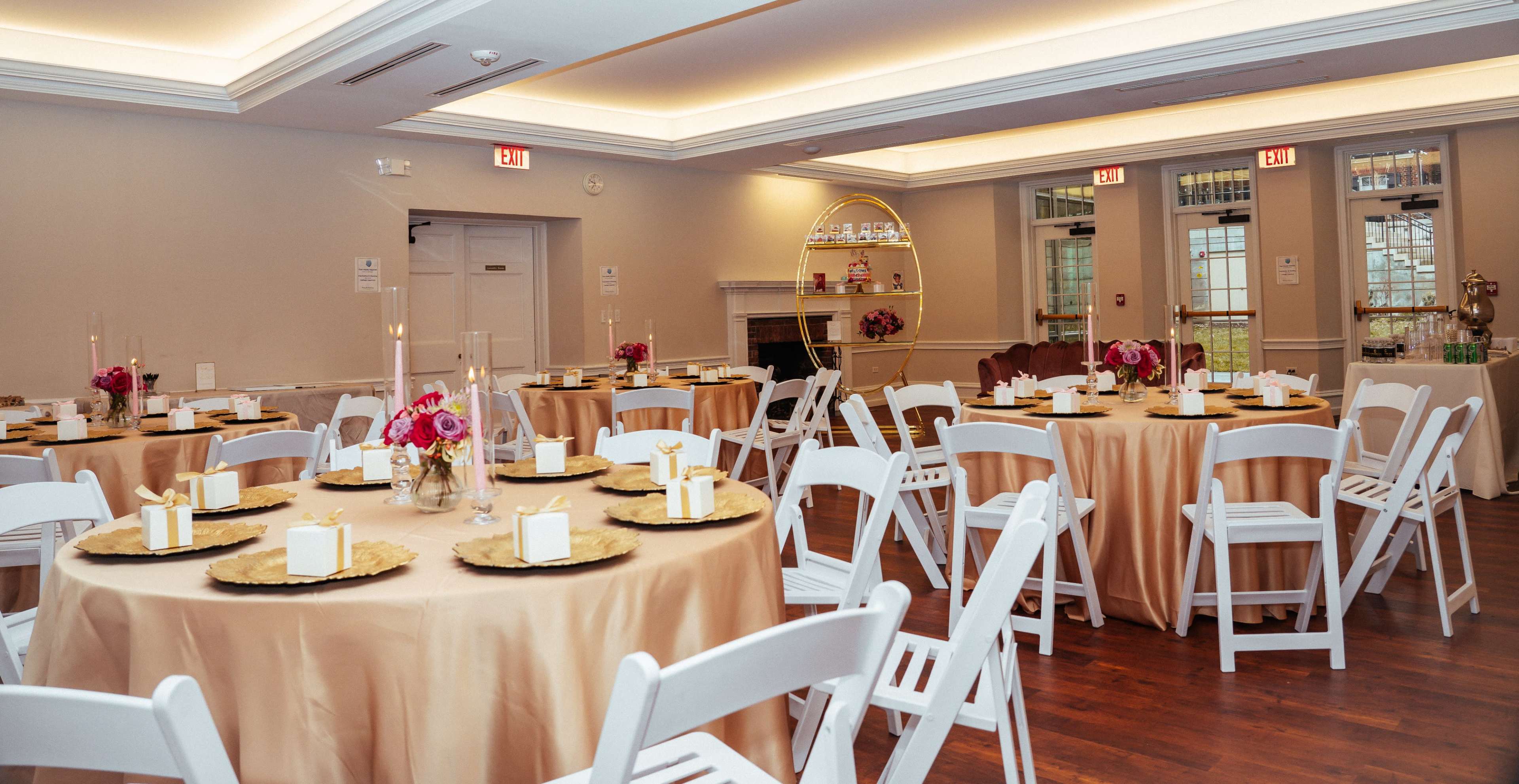 A banquet room set with round tables covered in gold and beige linens, each adorned with floral centerpieces and white chairs arranged around them.