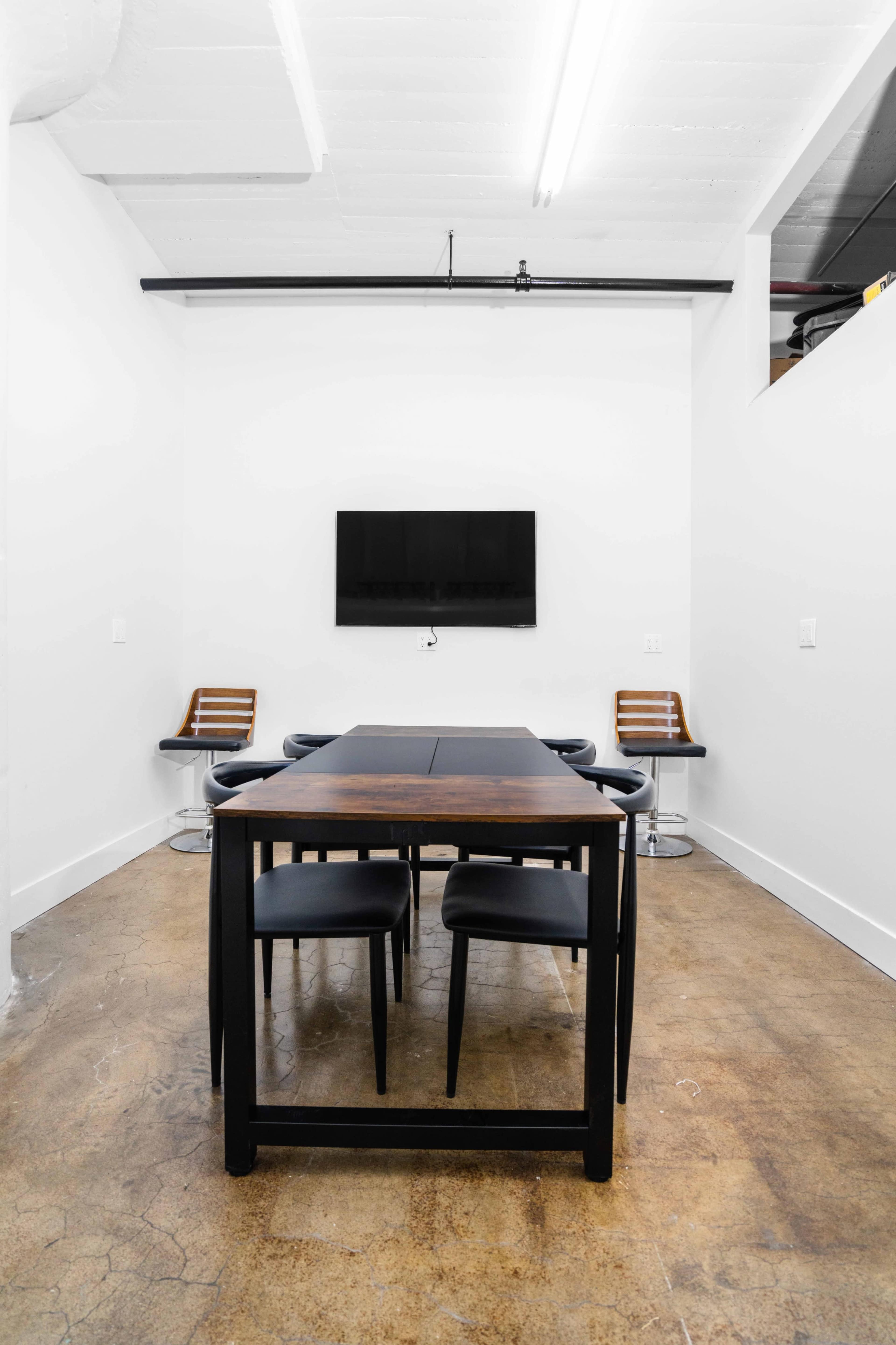 A minimalist meeting room with a black table and chairs, a wall-mounted TV, and concrete flooring.