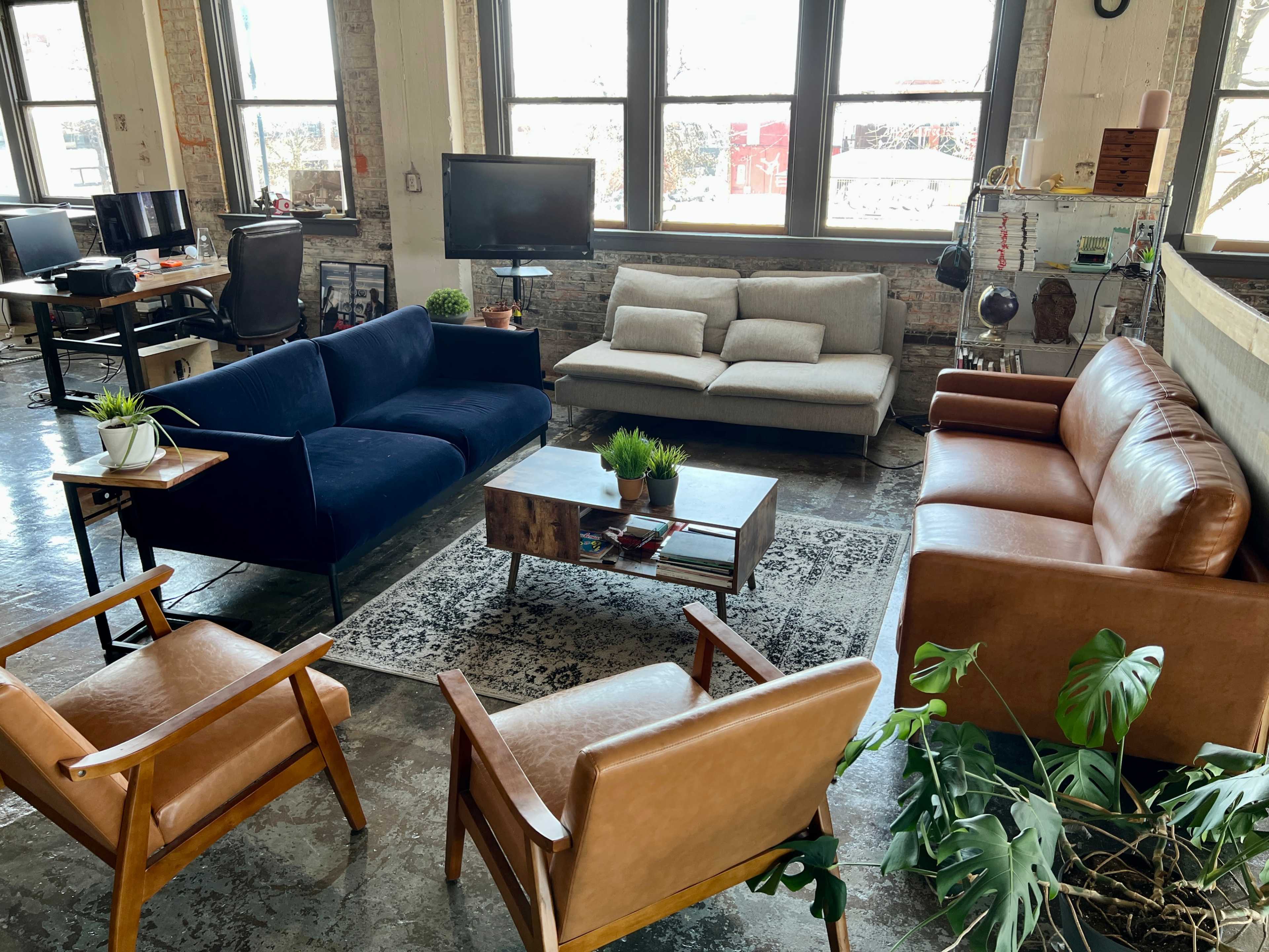The image shows a modern lounge area with various couches and chairs arranged around a coffee table, surrounded by large windows that let in natural light.