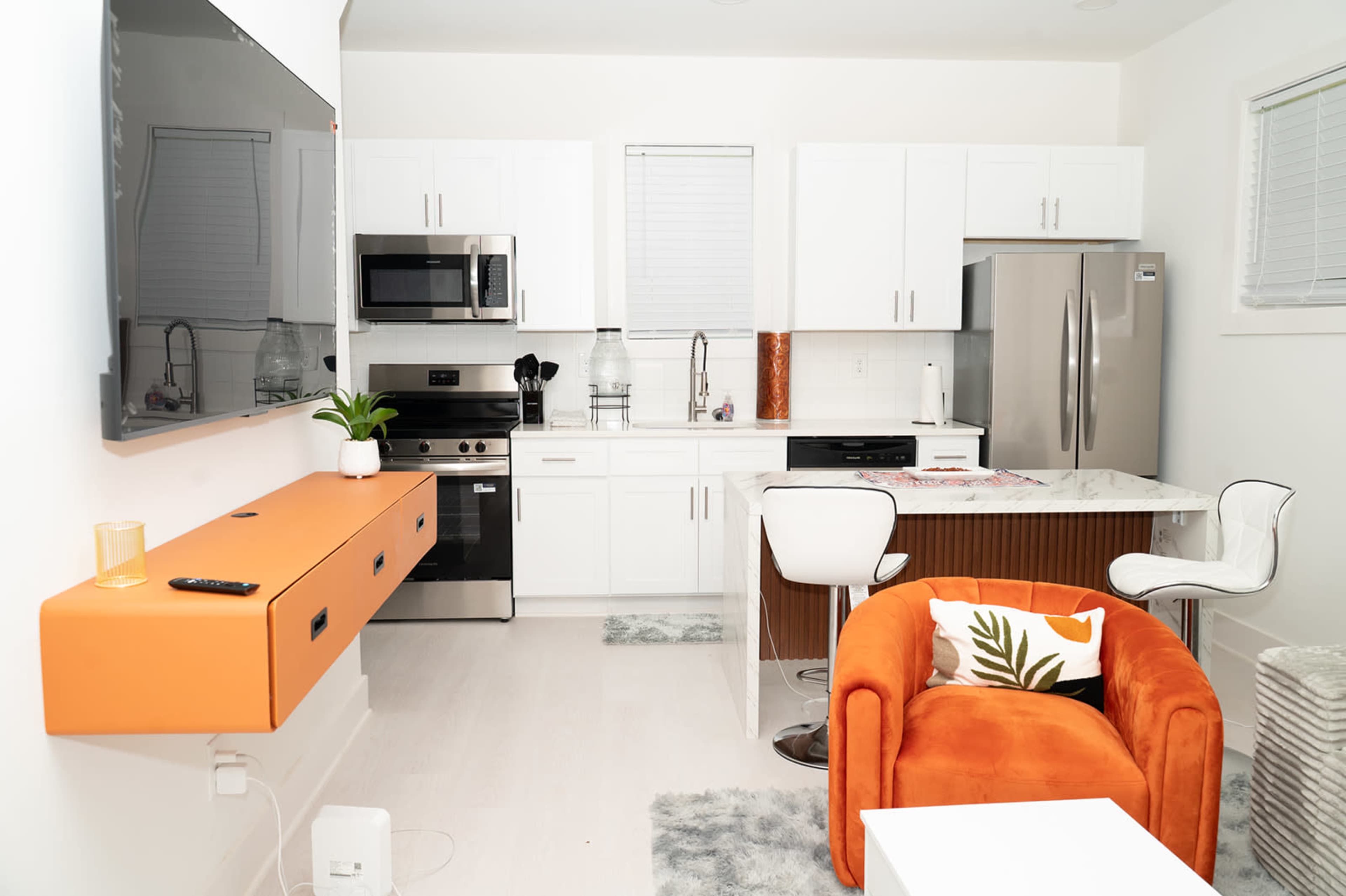 The image shows a modern kitchen and living area featuring white cabinetry, stainless steel appliances, an orange console table, and a cozy orange chair.
