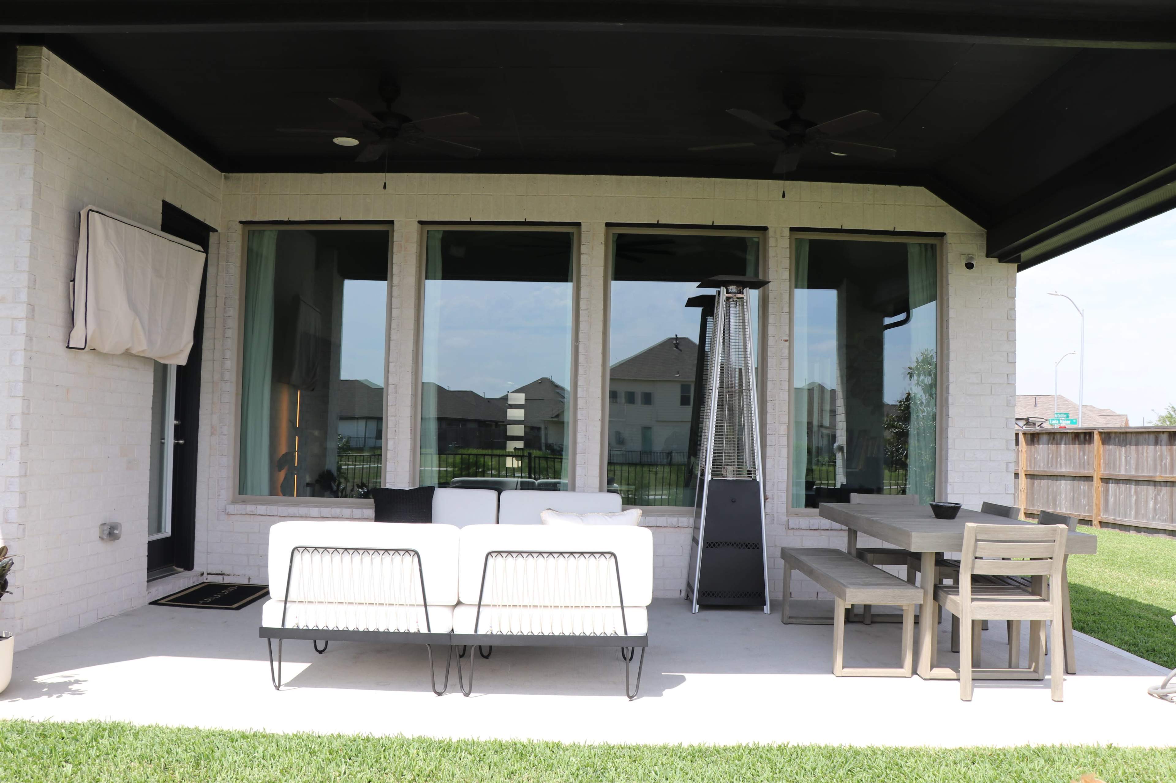 The image shows a covered patio featuring a white sofa, a dining table with chairs, and a tall patio heater next to large windows overlooking a lawn.