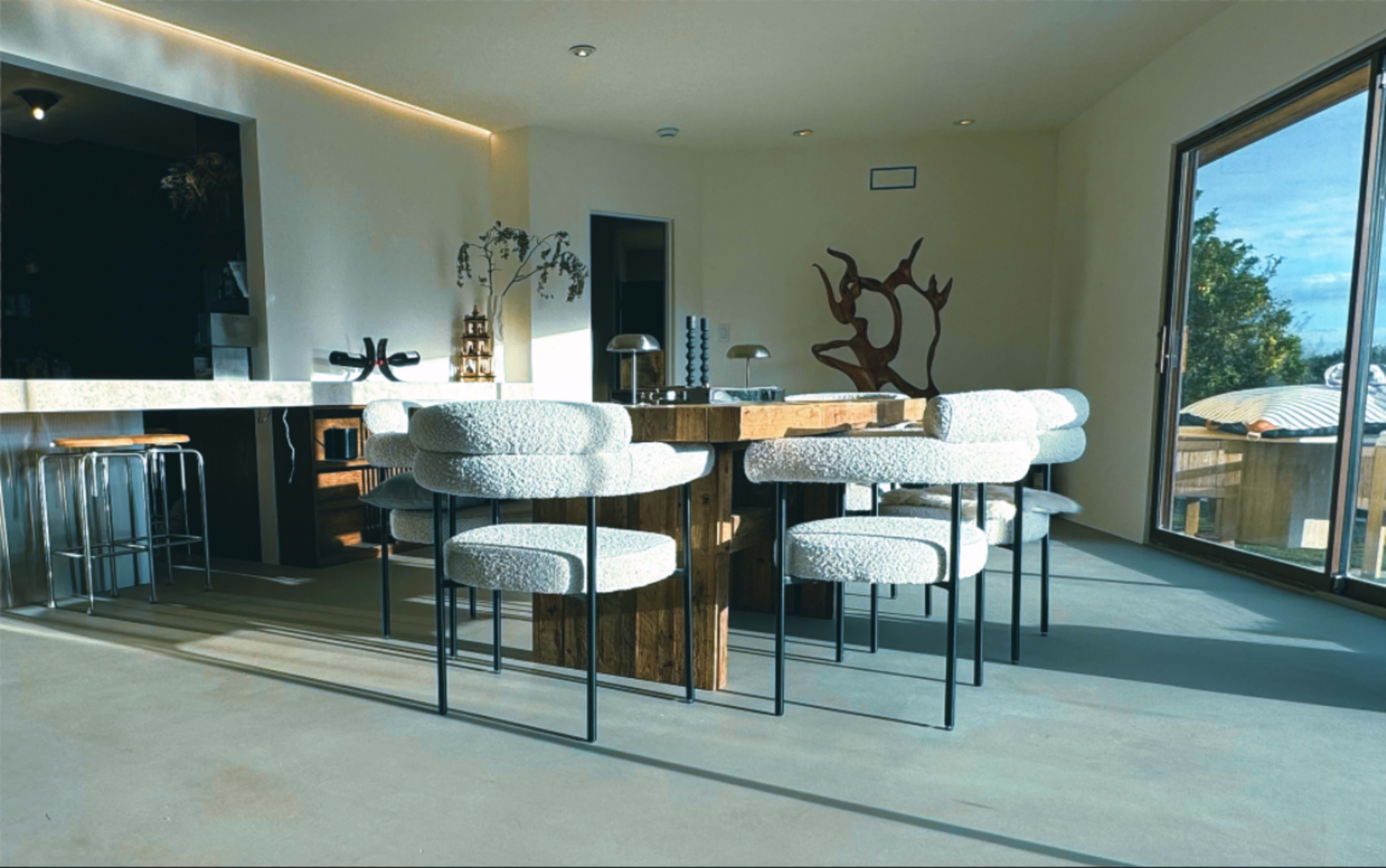 The image shows a modern dining area with a large wooden table surrounded by upholstered chairs and a large window letting in natural light.