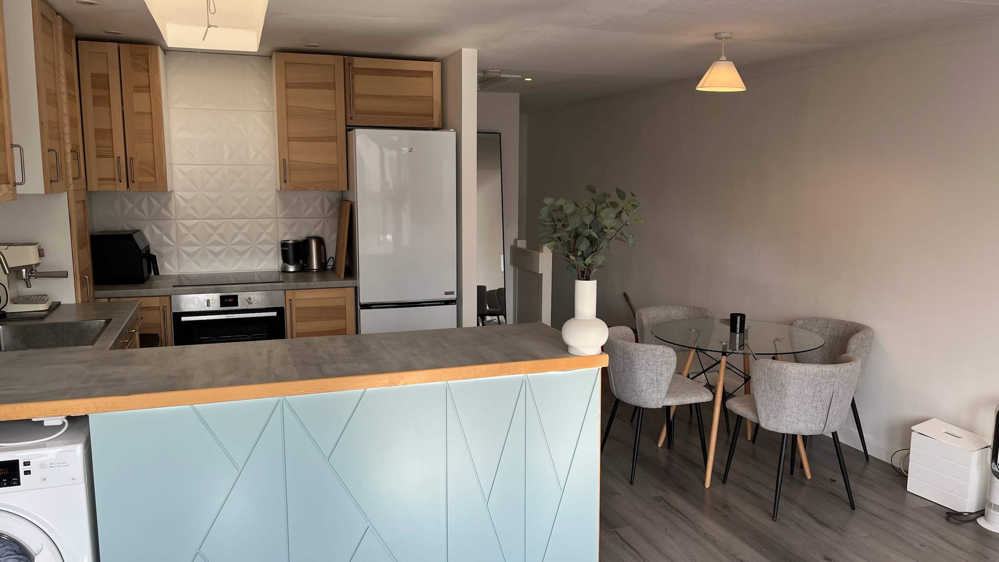 The image shows a modern kitchen with wooden cabinets and a dining area featuring a glass table and four chairs.