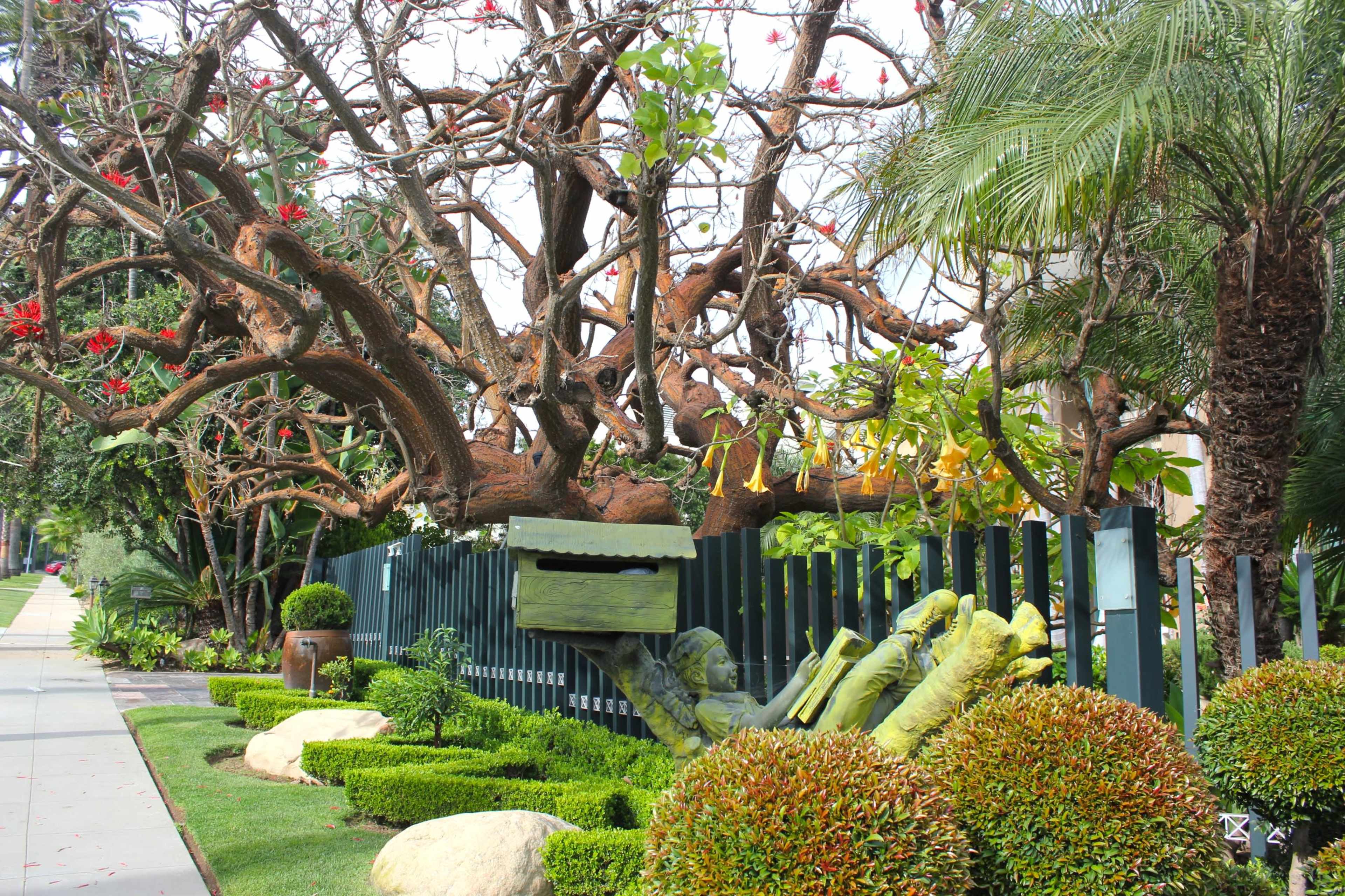A large tree with vibrant flowers stands beside a manicured lawn, featuring a unique sculpture of a hand partially protruding from the ground.