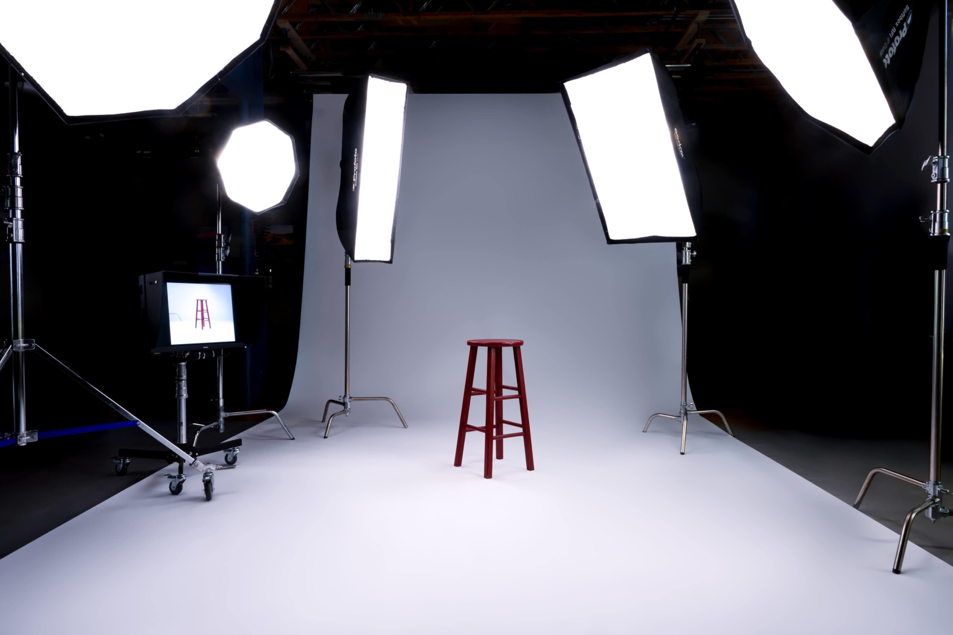 A red stool is positioned in the center of a photography studio, surrounded by large softbox lights and a gray backdrop.