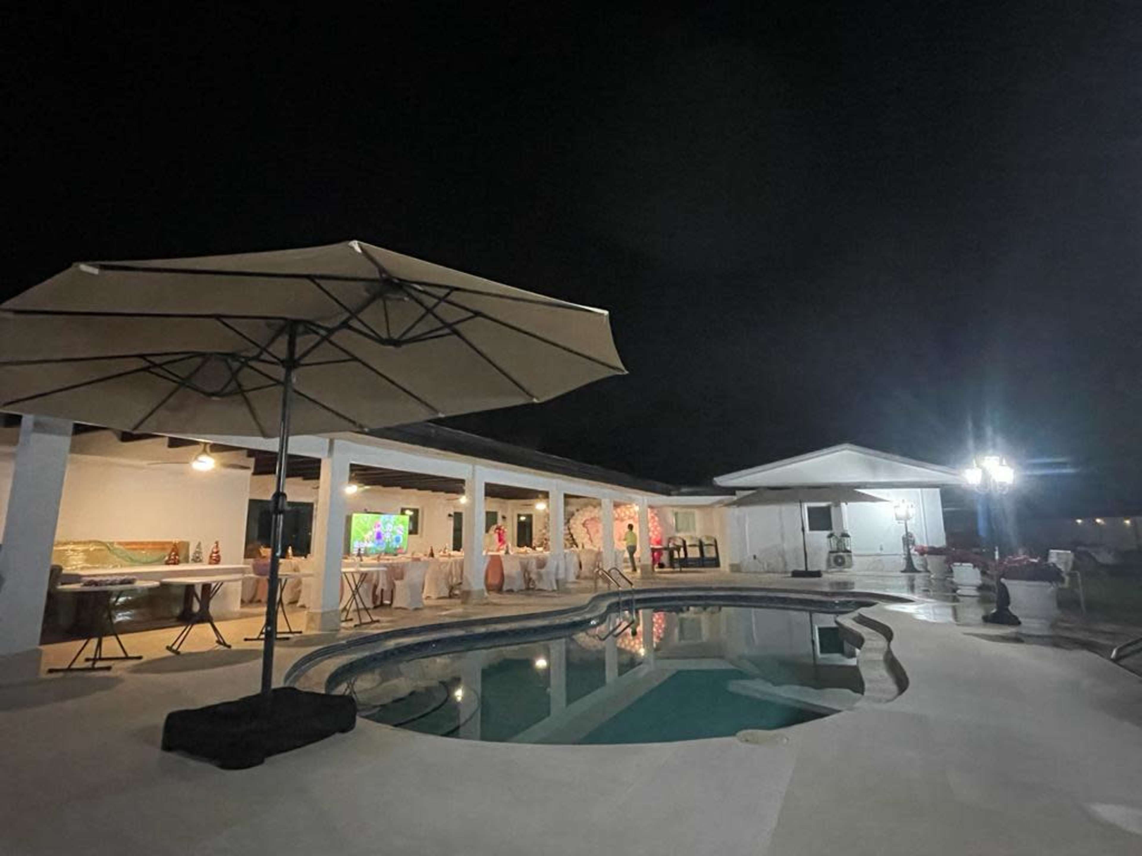 A well-lit backyard features a swimming pool surrounded by a patio, with a large umbrella and a house in the background at night.